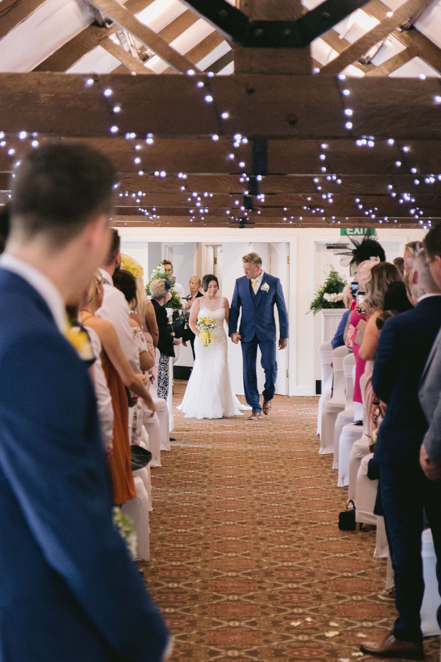 Couple walk down aisle as newlyweds; guests line the room under twinkling lights in a warm, rustic setting.