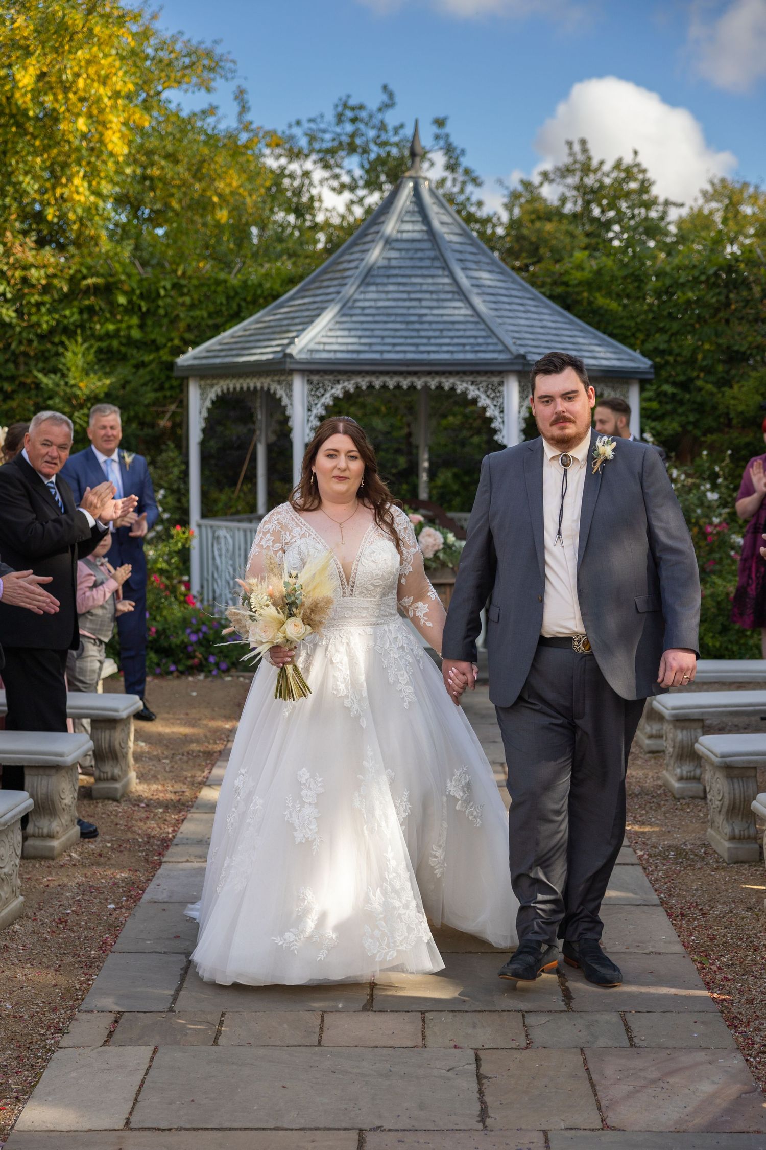 Couple walk down aisle at Bowburn Hall; gazebo backdrop and guests applauding in garden setting.