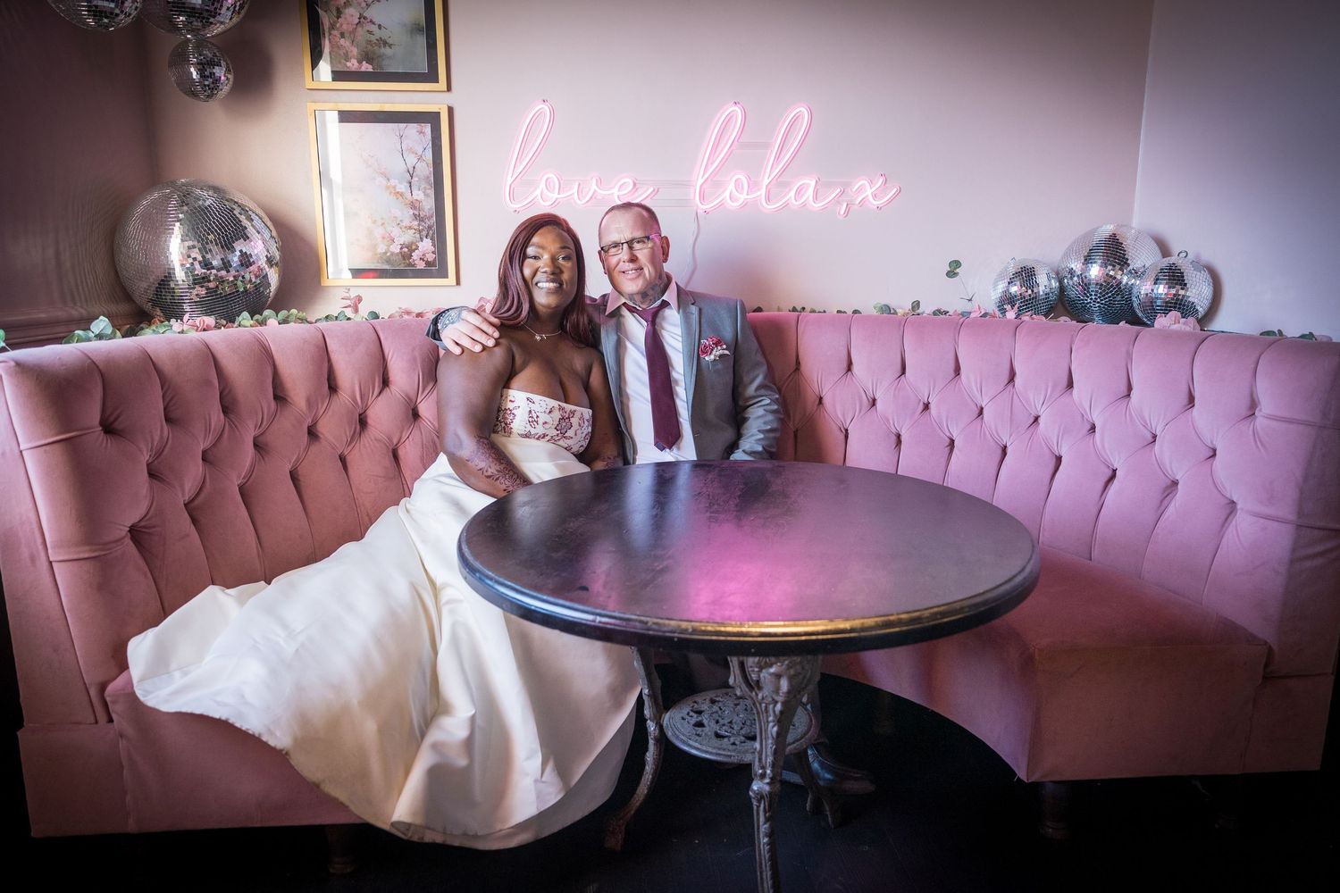 Bride and groom sit in pink booth under “love” sign at Tynemouth venue, smiling in a cozy, stylish setting.