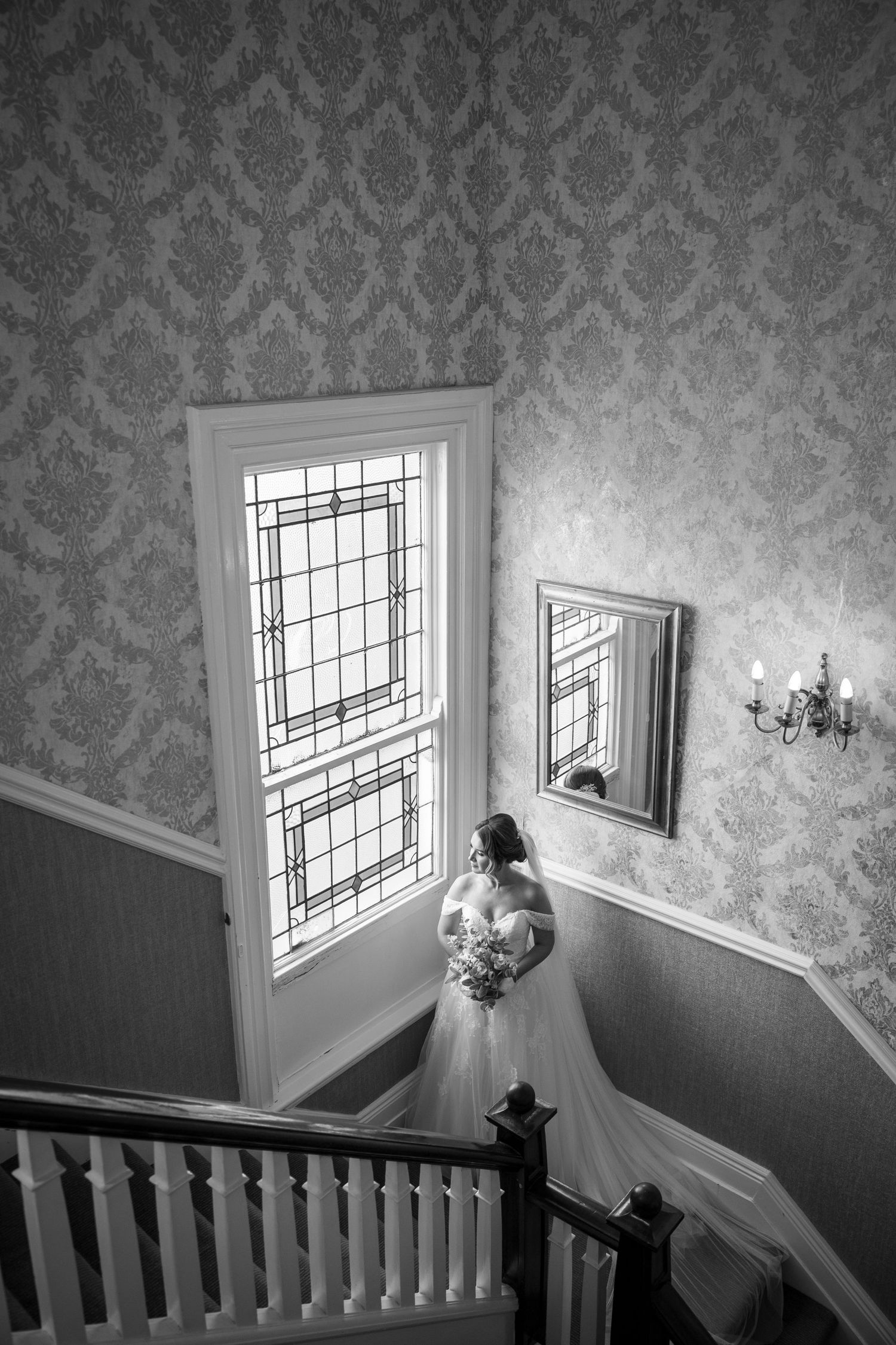 Bride pauses on staircase, bouquet in hand; black-and-white shot with patterned walls and soft window light.