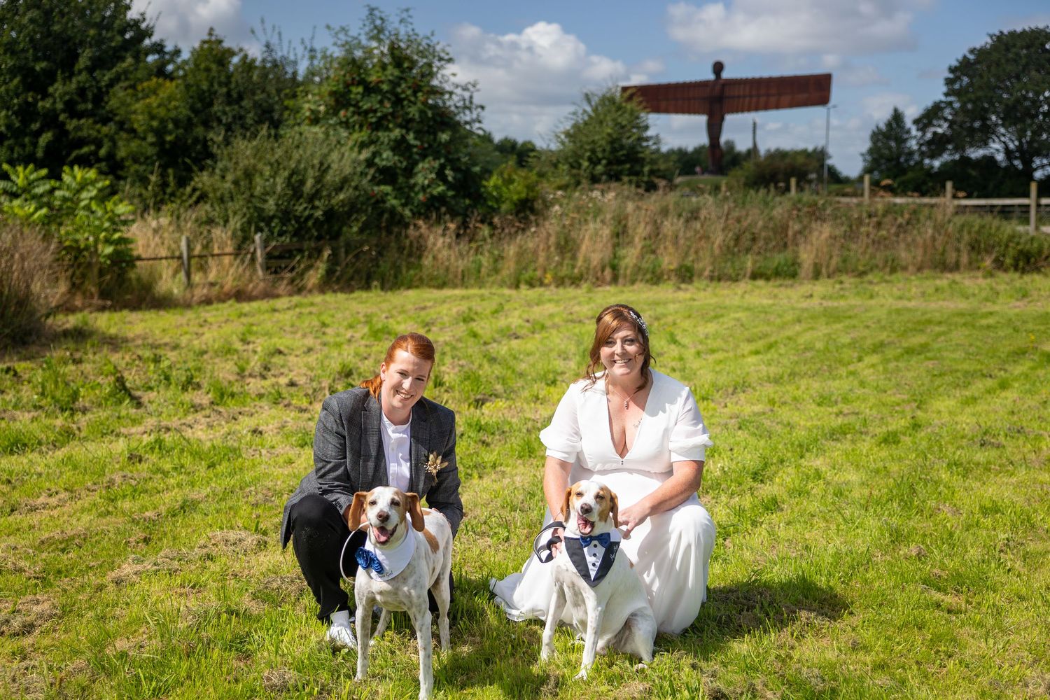 Same-sex couple with dogs in sunny field; relaxed, joyful portrait from a low angle view.