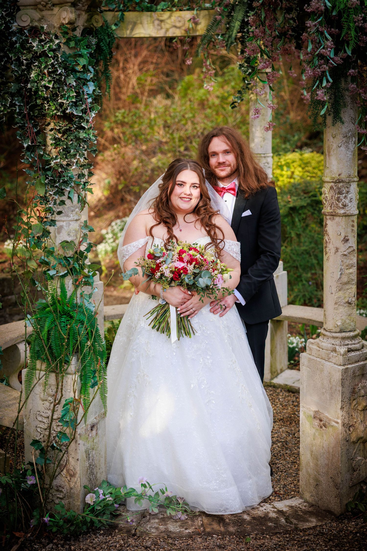 Bride and groom pose under ivy arch at Whitworth Hall; lush garden setting with romantic feel.