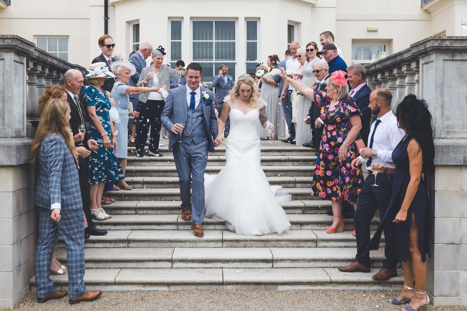 Bride and groom walk down steps at Seaham Hall, confetti moment with guests celebrating, joyful wedding day scene outdoors.