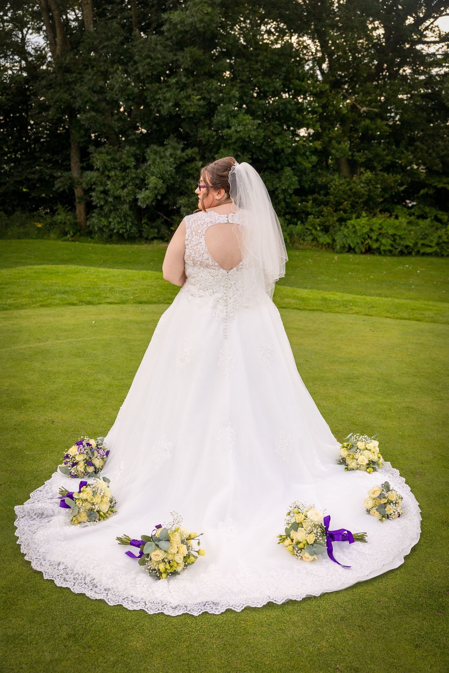Bride stands on lawn with bouquet details arranged around her train; elegant, creative portrait in a garden setting.