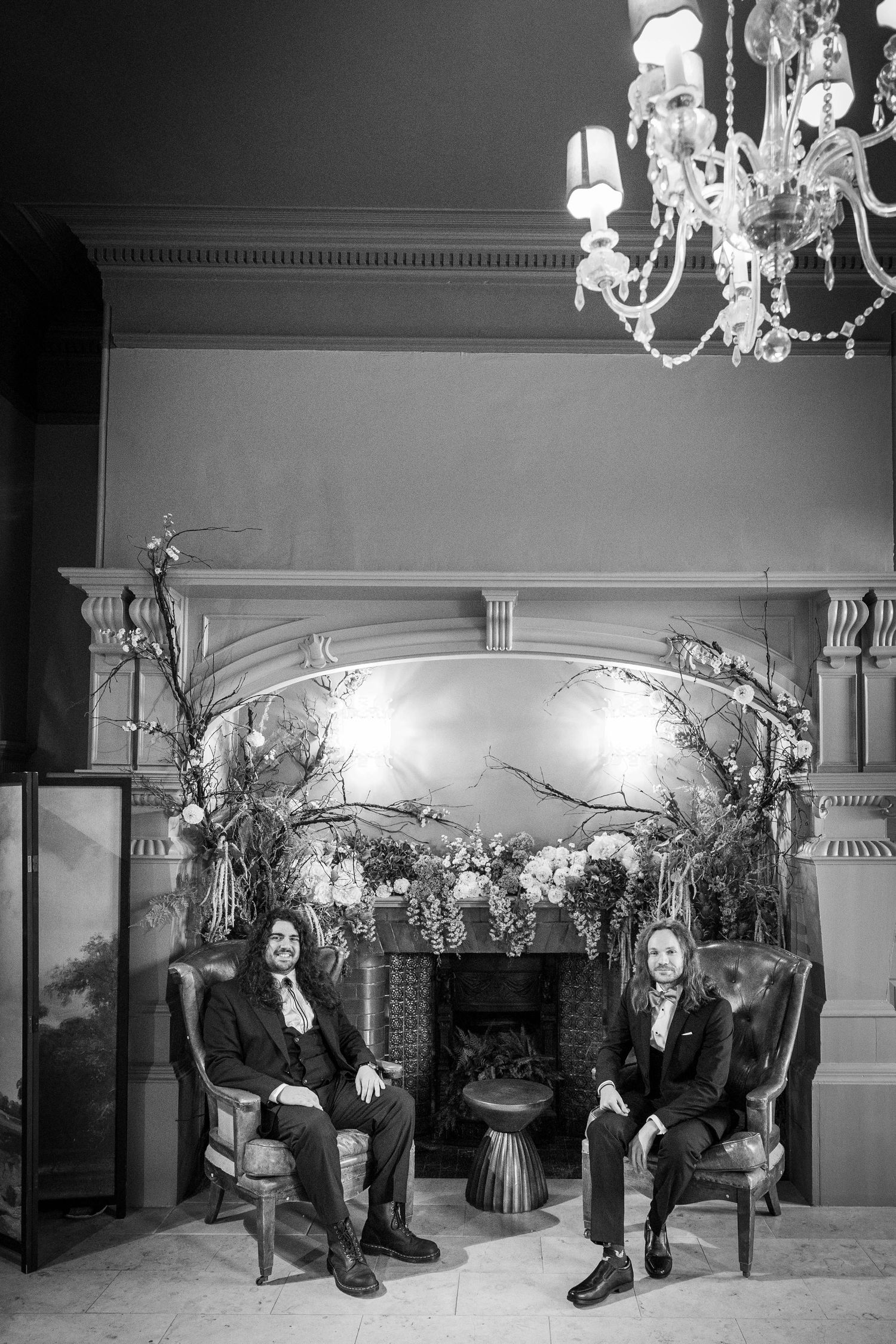 Grooms sit in armchairs at Whitworth Hall; elegant black-and-white portrait with chandelier and floral backdrop.