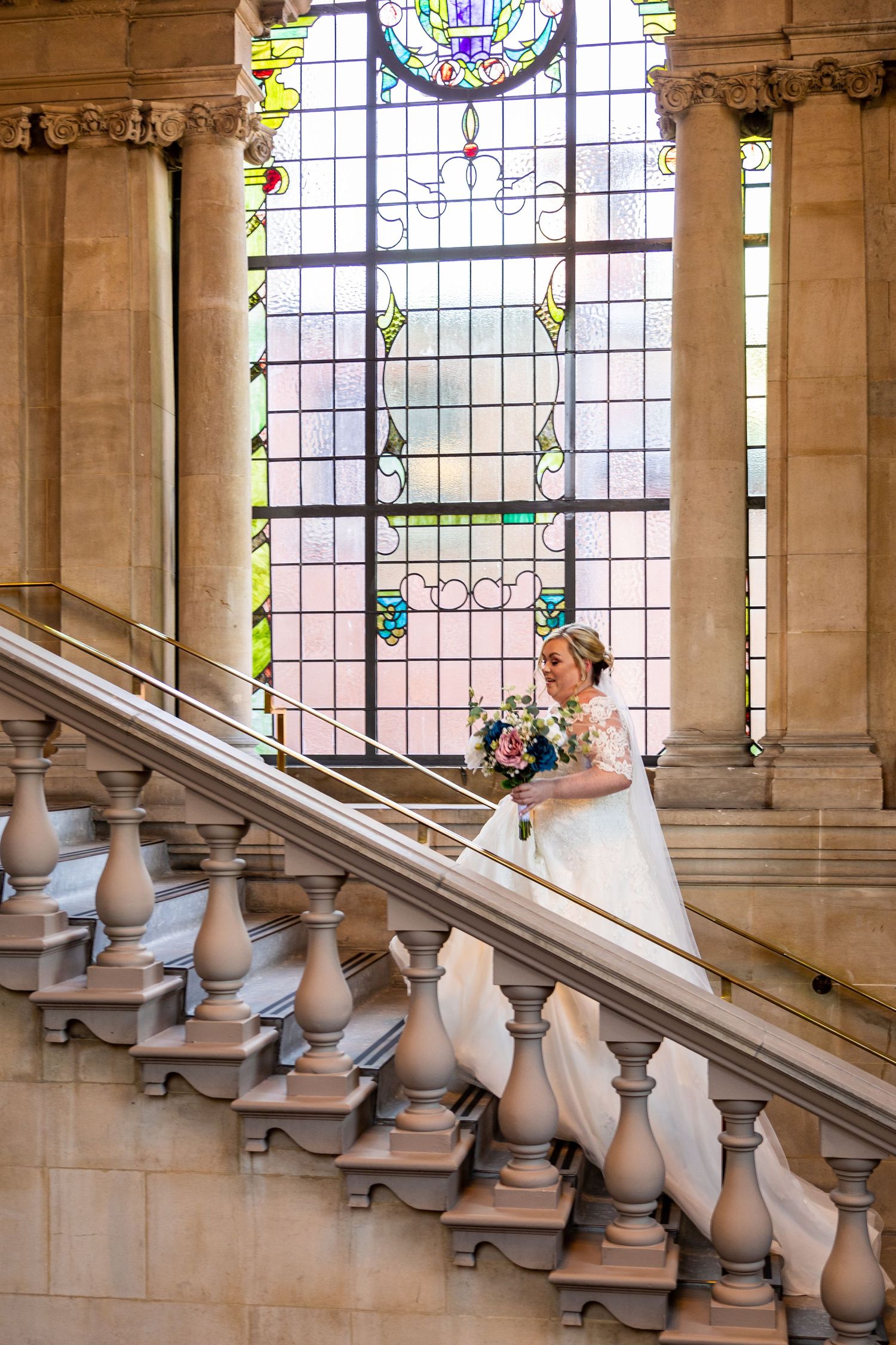 Bride ascends staircase at South Shields Registry Office; stained glass backdrop adds elegance.