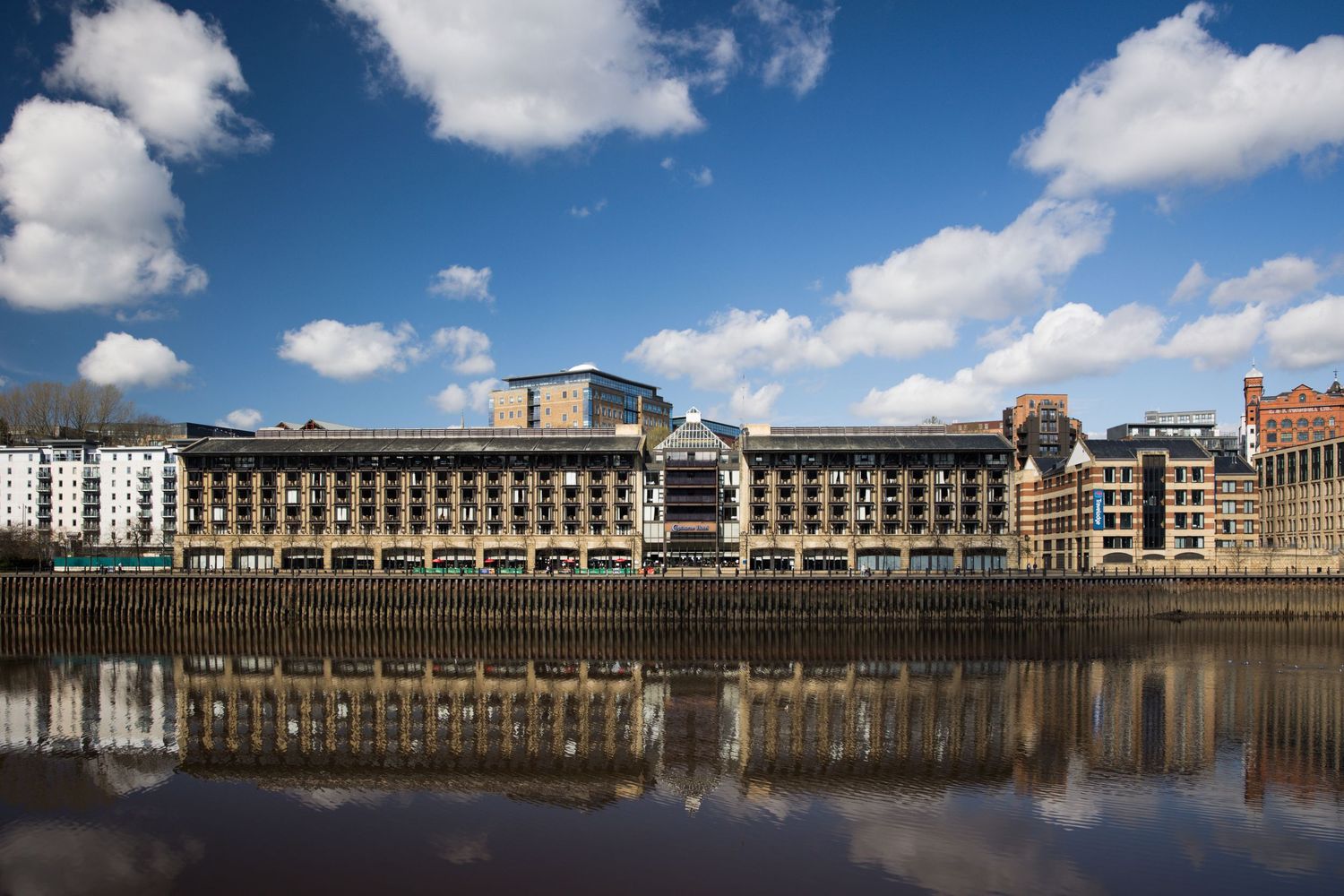 Riverside building reflected in calm water under blue sky; modern architecture along the waterfront.