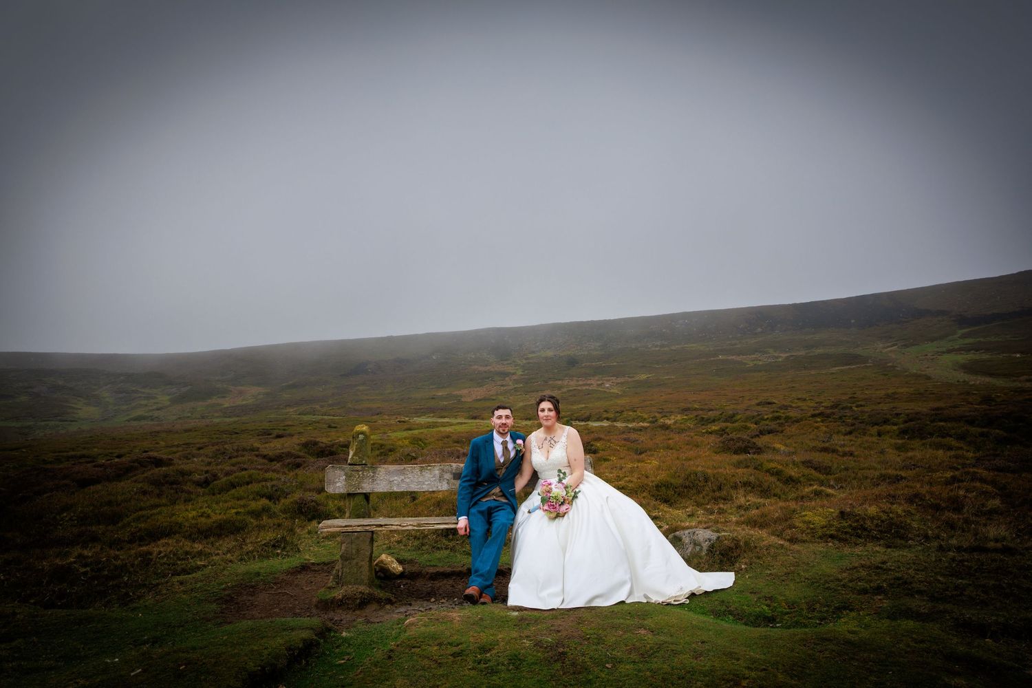 Couple seated on bench at Danby Castle, overlooking Yorkshire Moors; serene, windswept landscape backdrop.
