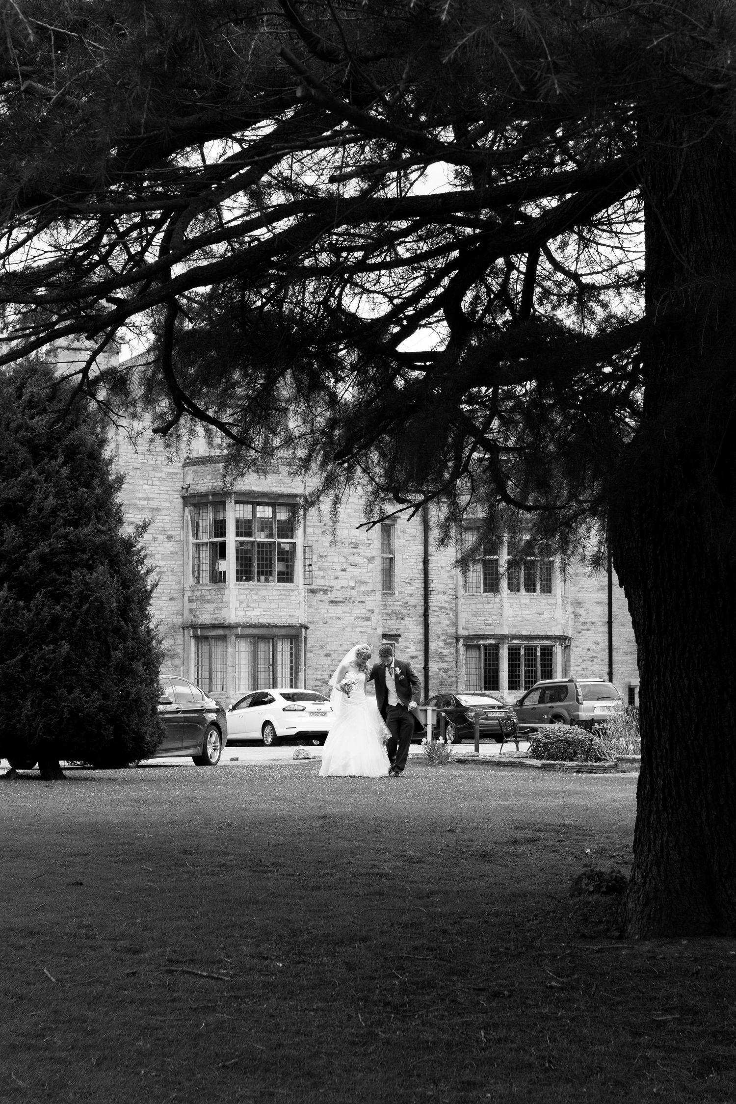 Bride and groom walk beneath trees at Redworth Hall Hotel, elegant black and white wedding portrait in scenic grounds