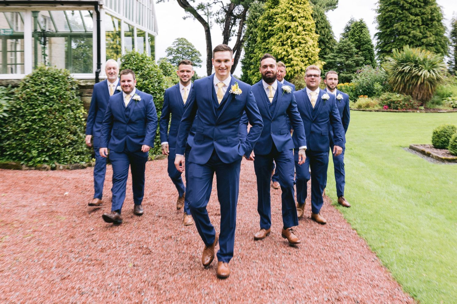 Groom and groomsmen walking confidently through gardens in matching blue suits on wedding day