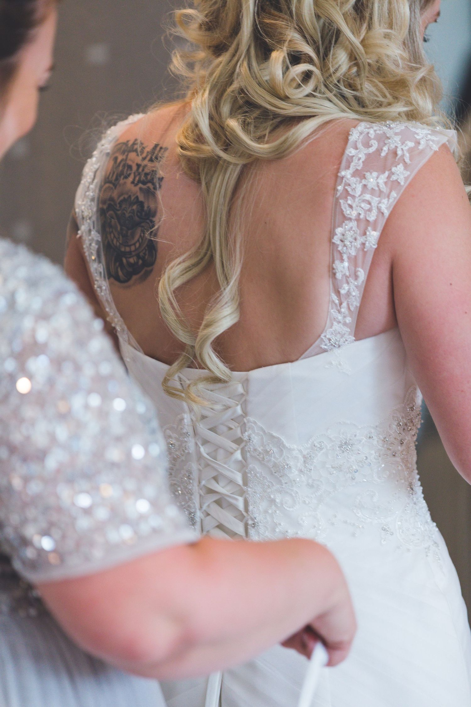 Bride getting ready, lace wedding dress detail with corset back, bridesmaid helping, elegant bridal preparation moment.