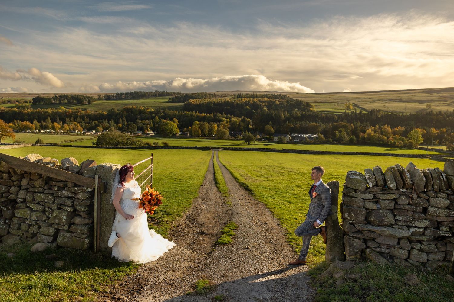 Golden-hour countryside portrait in Wolsingham, with a bride and groom framed by stone walls and a winding rural track across rolling fields