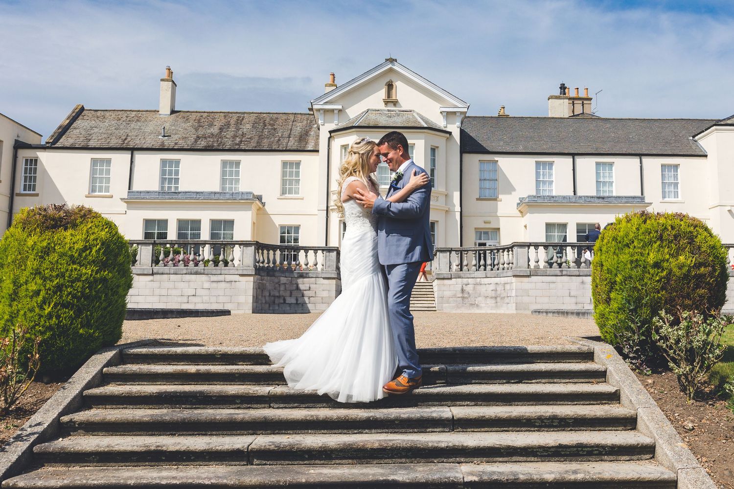 Couple embrace on steps at Seaham Hall; elegant white facade and sunny garden setting.