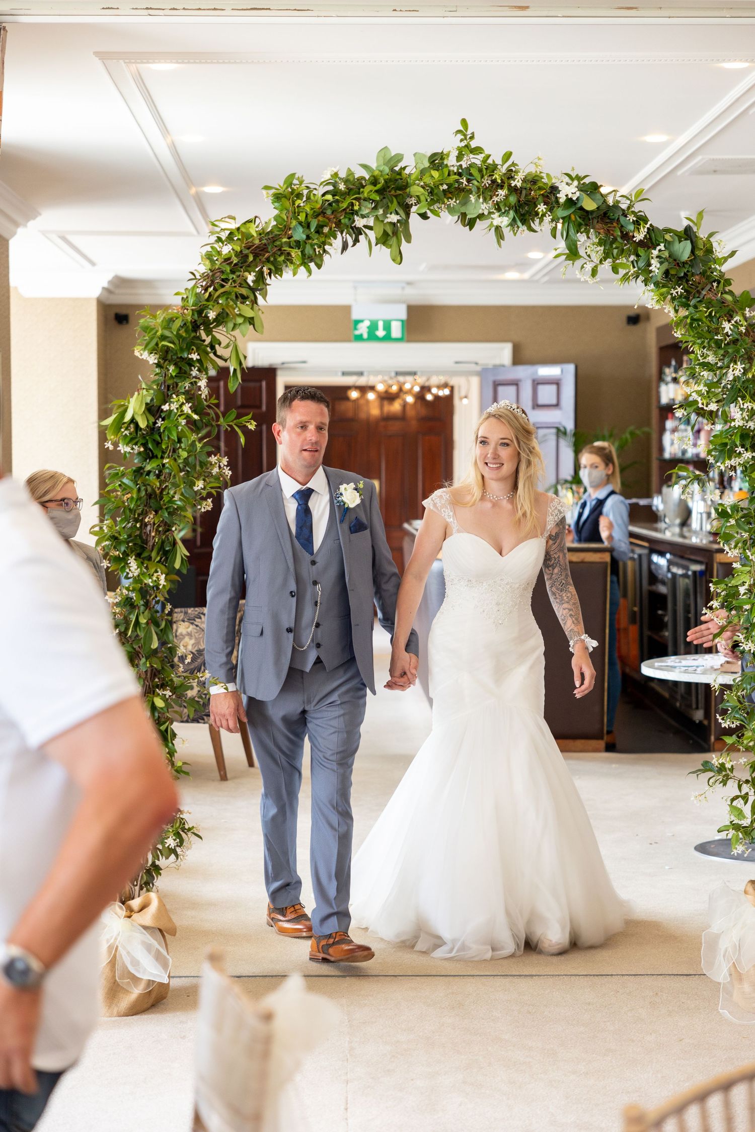 Bride and groom walk under floral arch at Seaham Hall, indoor wedding ceremony entrance, romantic aisle moment.