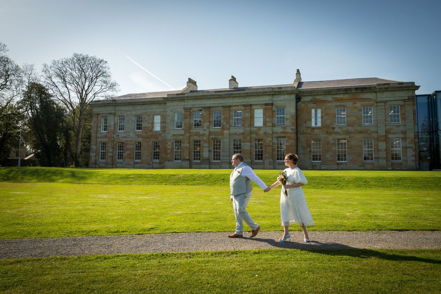 Couple walks hand in hand at The Story, Durham; grand historic building and sunny lawn backdrop.