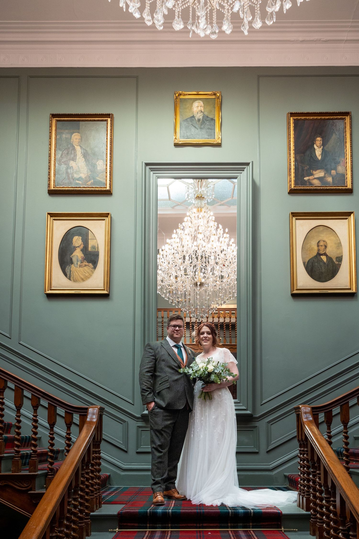Couple pose on staircase beneath chandelier; elegant portraits and classic interior setting.