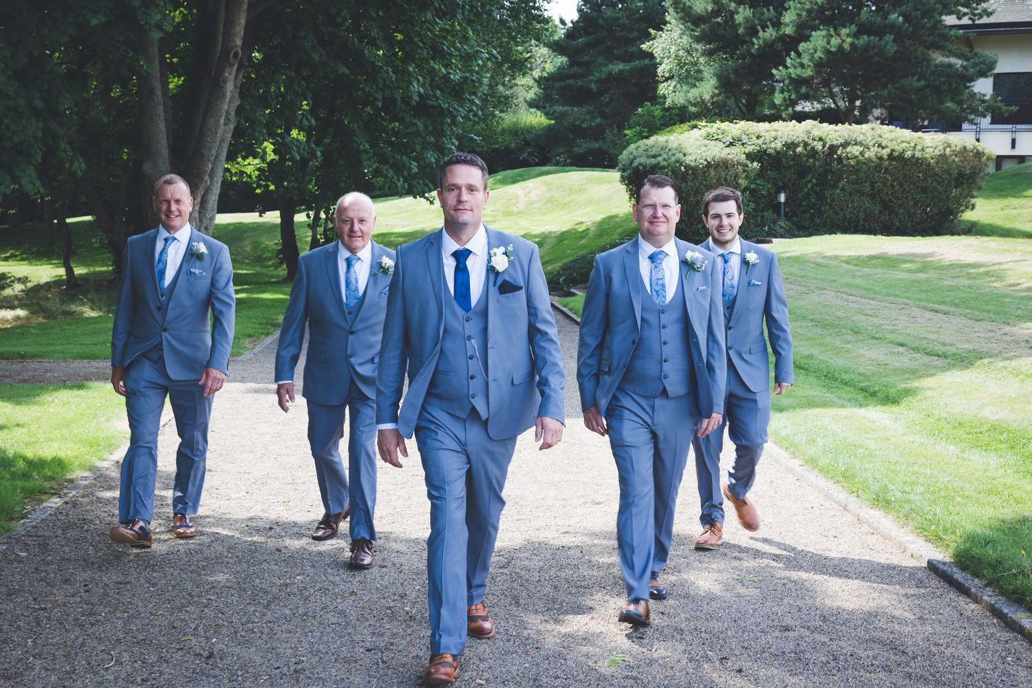 Groom and groomsmen walking at Seaham Hall, stylish blue suits, outdoor wedding party portrait in elegant garden setting.