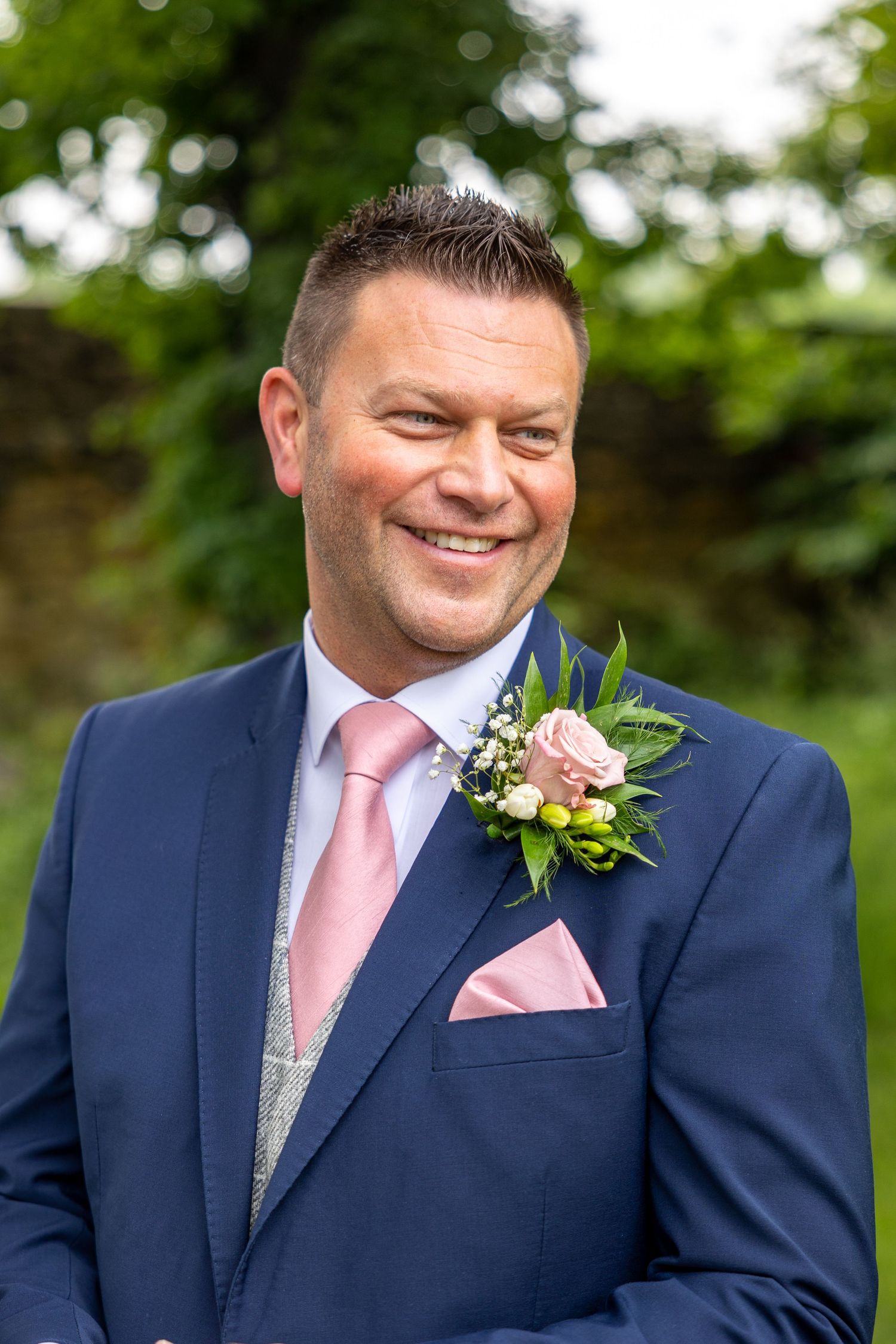 Smiling groom in navy suit with pink tie and rose boutonniere, photographed outdoors with soft greenery background