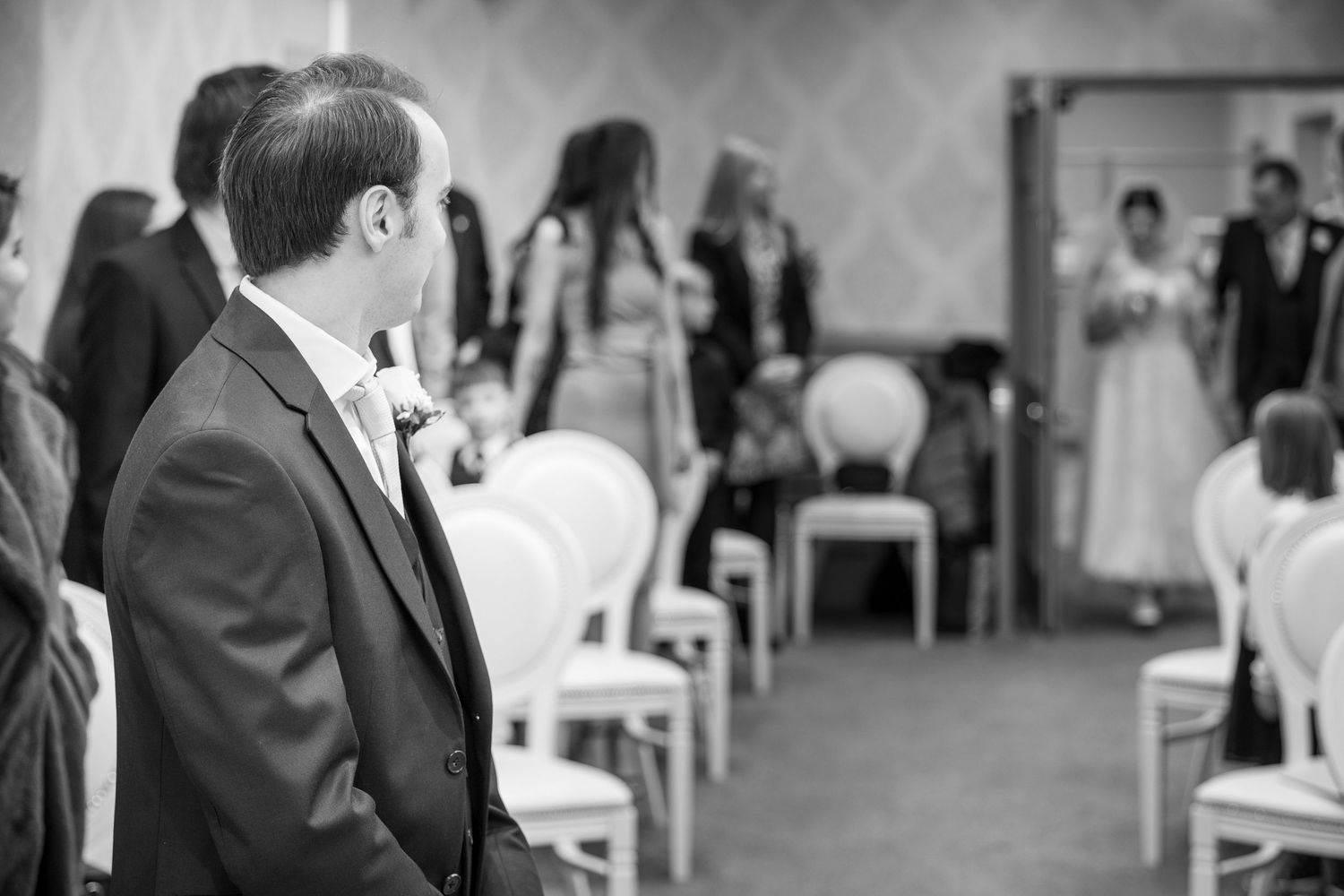 Groom waits at aisle, glancing back as bride enters; guests seated, soft focus, black-and-white ceremony moment.