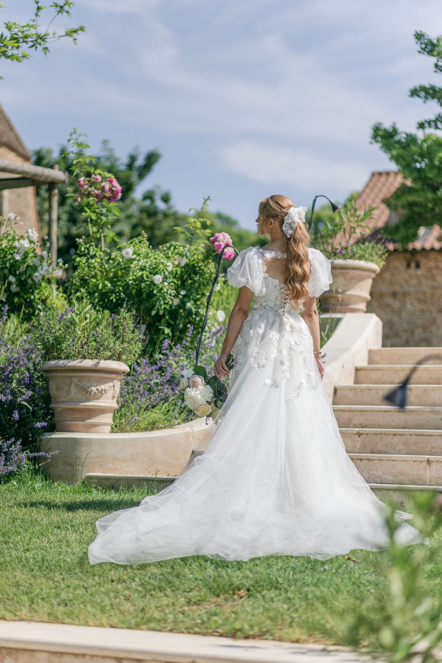 A bride in a flowing white gown poses near garden steps surrounded by lavender plants.