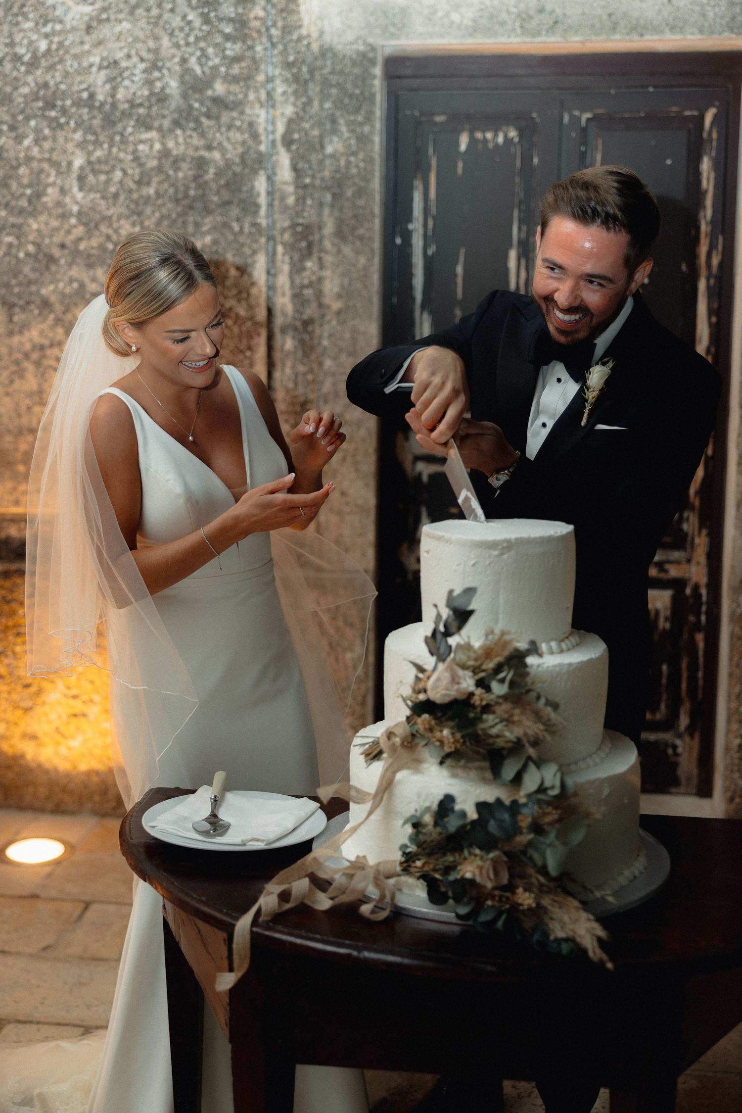 Newlyweds laugh while cutting their white wedding cake decorated with flowers.