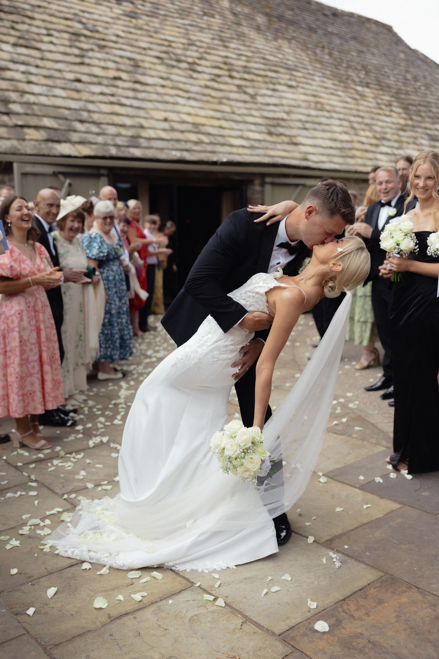 Wedding party dips in synchronized dance move outside stone building with confetti flying.