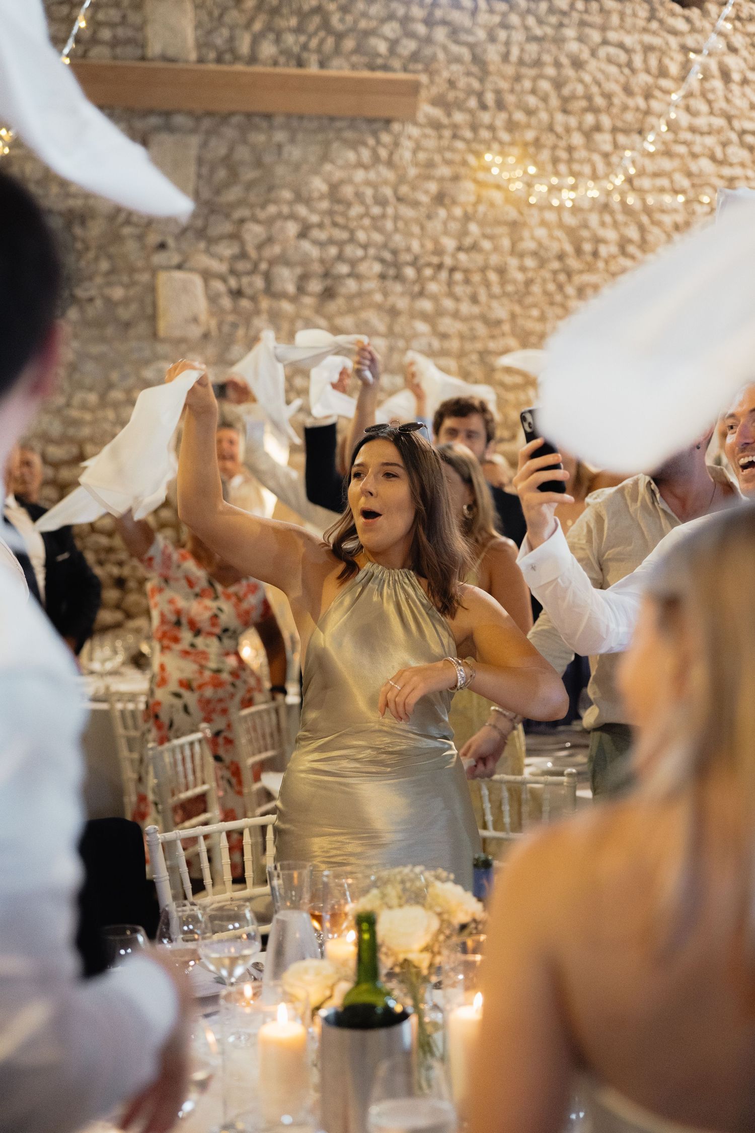 Wedding guests wave white napkins during celebration in rustic stone venue with candlelit tables.