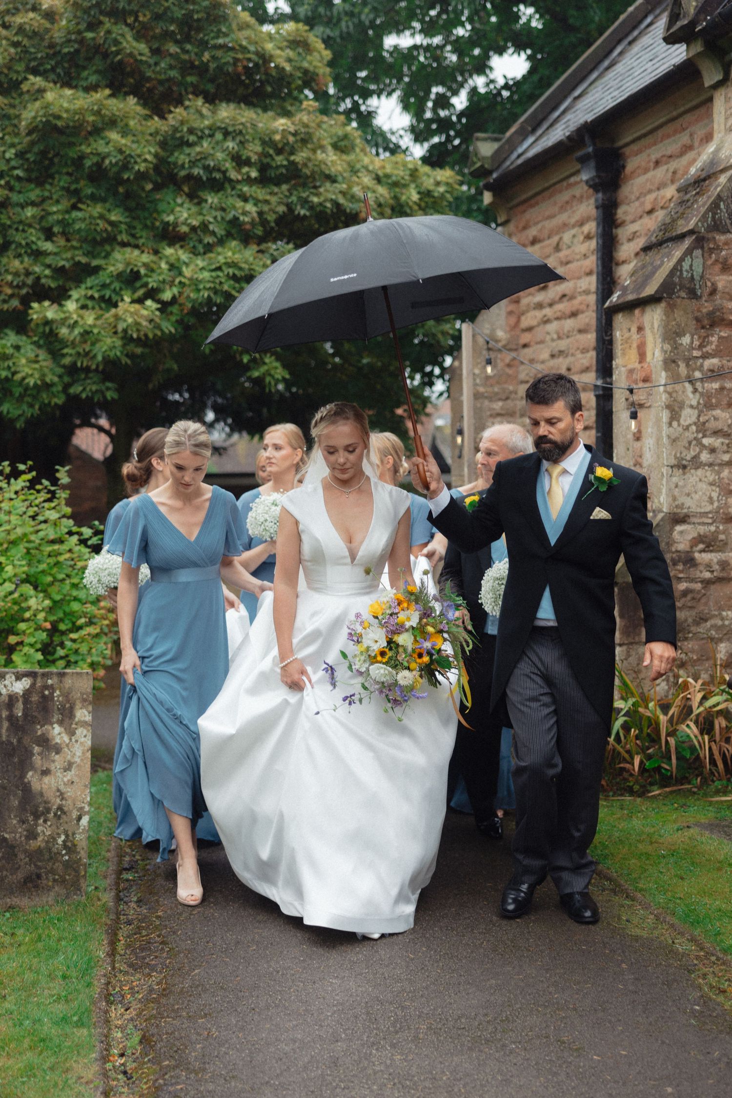 Wedding party walking along a stone path while holding umbrellas on a rainy day.
