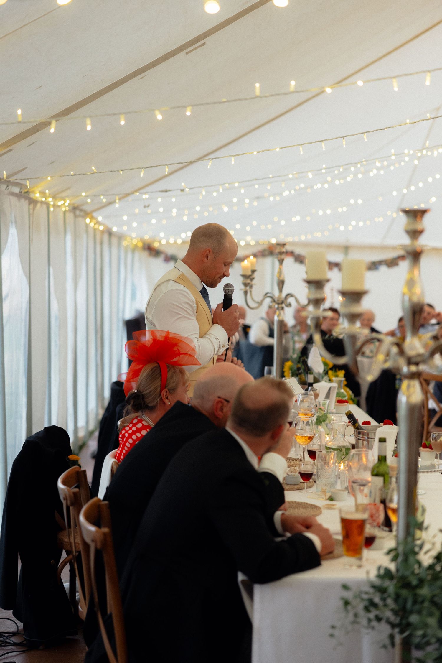 Guest delivers speech at wedding reception dinner under string lights.