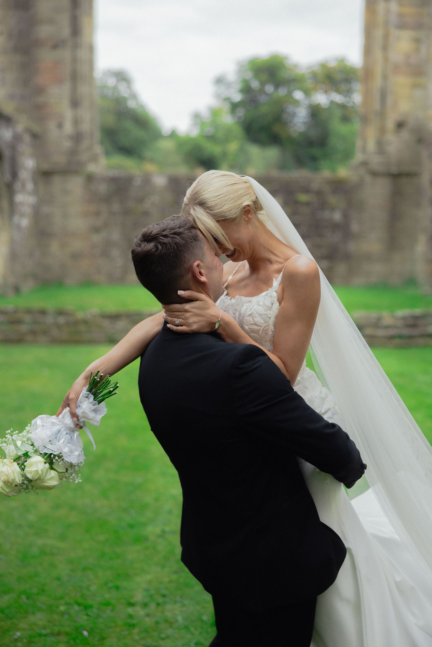 Romantic wedding couple embraces and dips against historic stone ruins and green landscape backdrop.