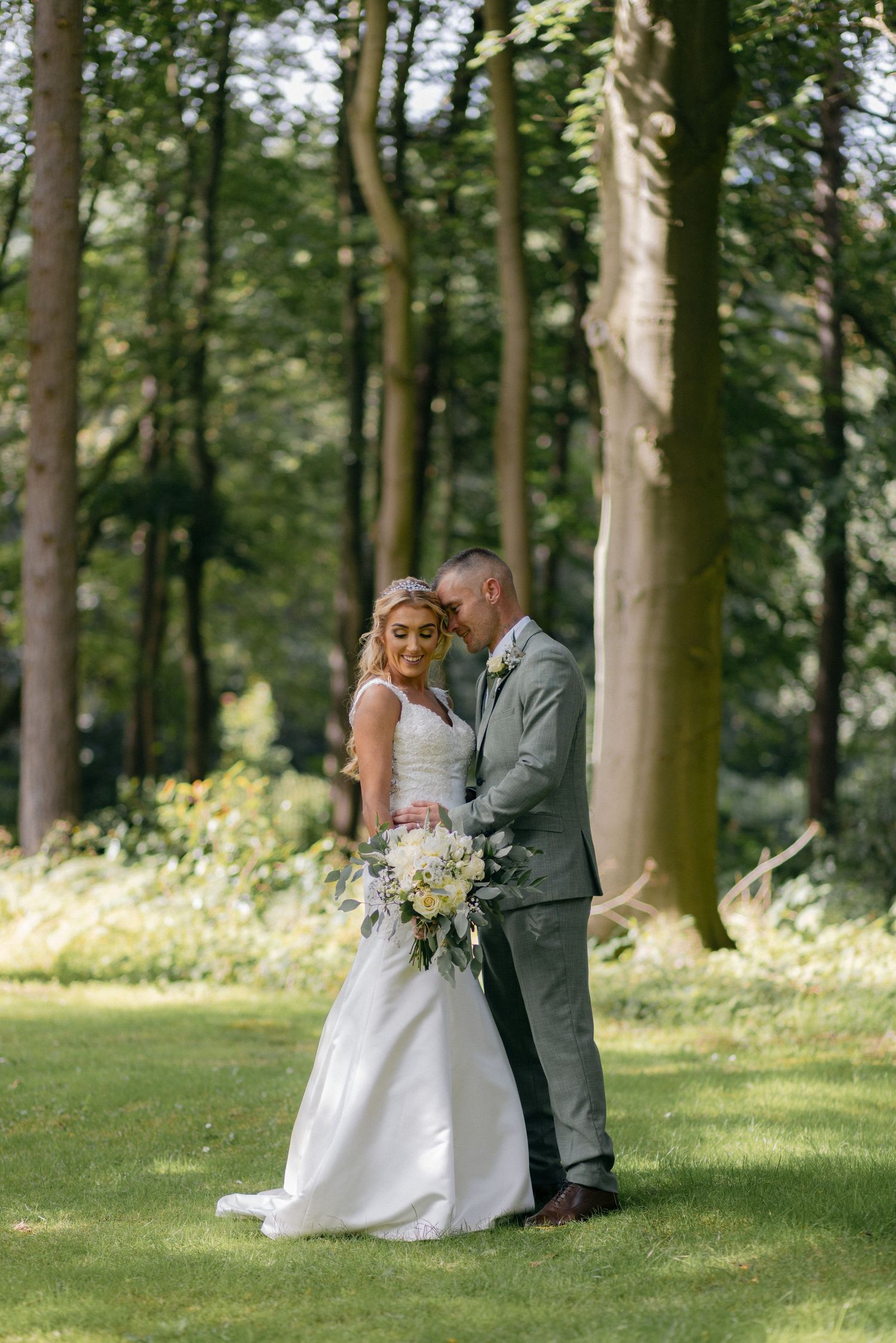 A tender wedding moment captured in a wooded garden setting with natural sunlight.