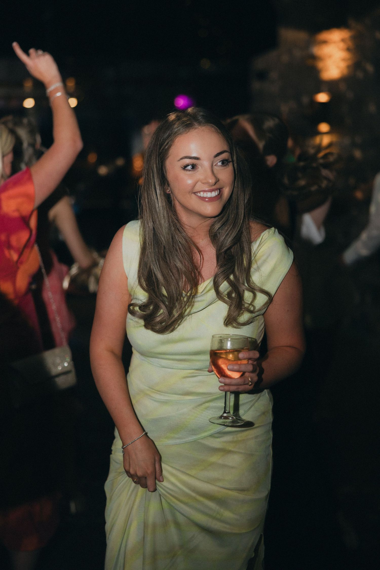 Guest in pale yellow dress enjoys celebration with wine glass.