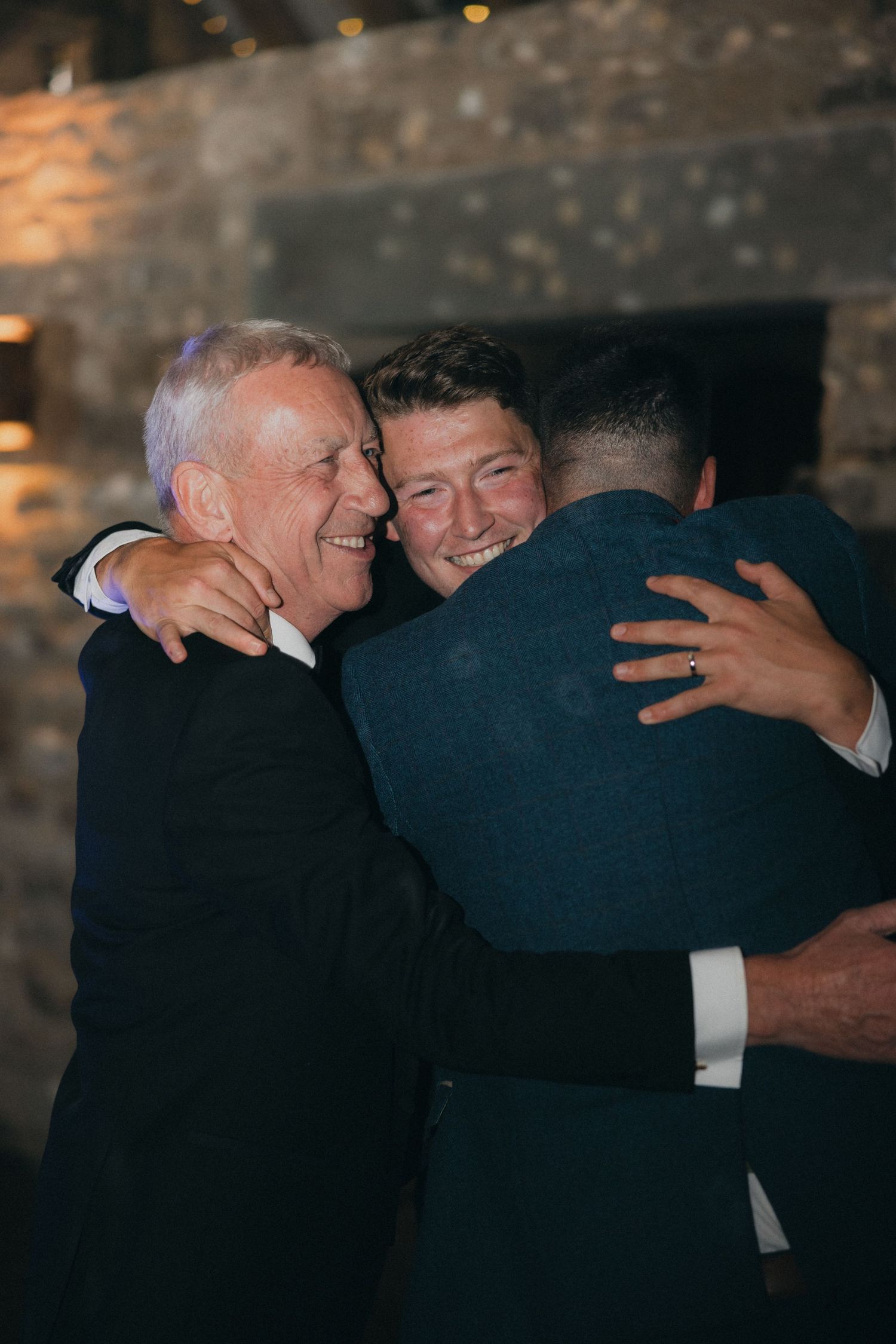 Three men share an emotional group hug at a formal event in a rustic venue.