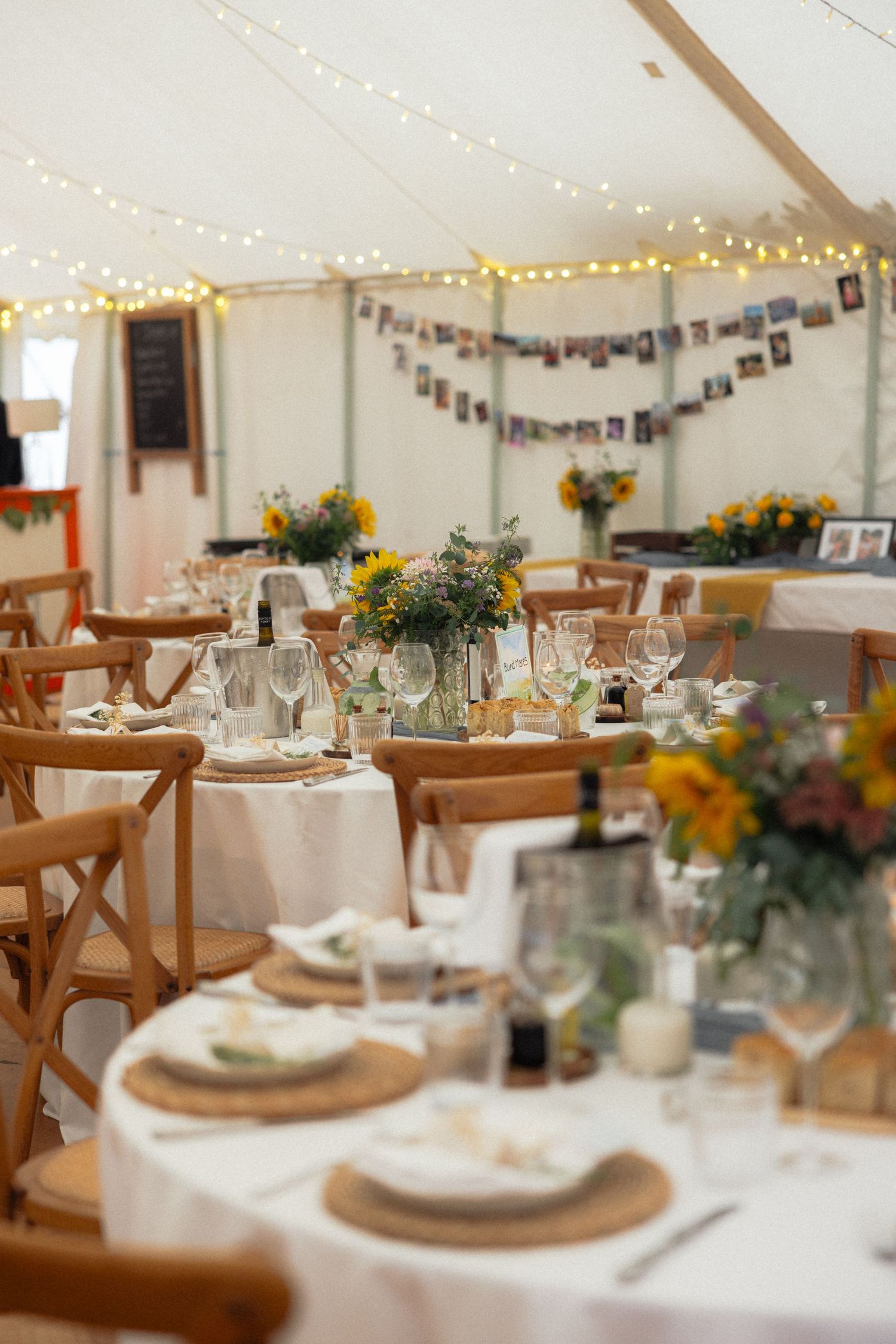 Wedding reception tent interior with elegant table settings, wooden chairs and string lights creating a romantic atmosphere.