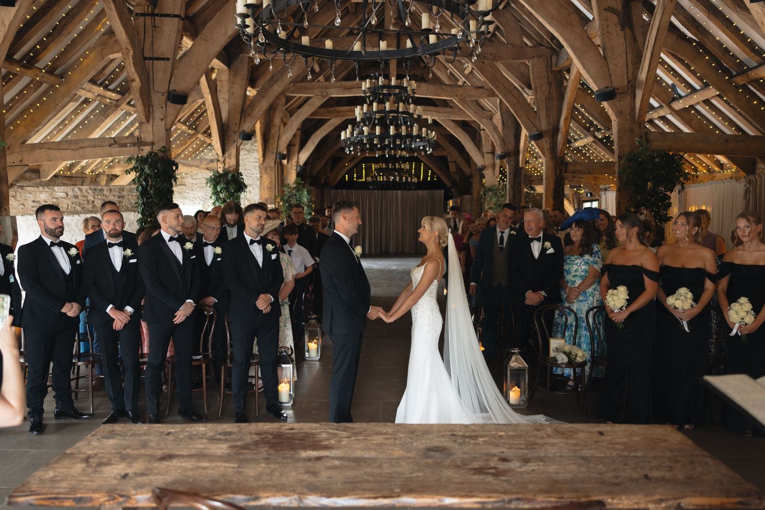 Wedding ceremony taking place in a rustic wooden barn venue with exposed beams and chandeliers.