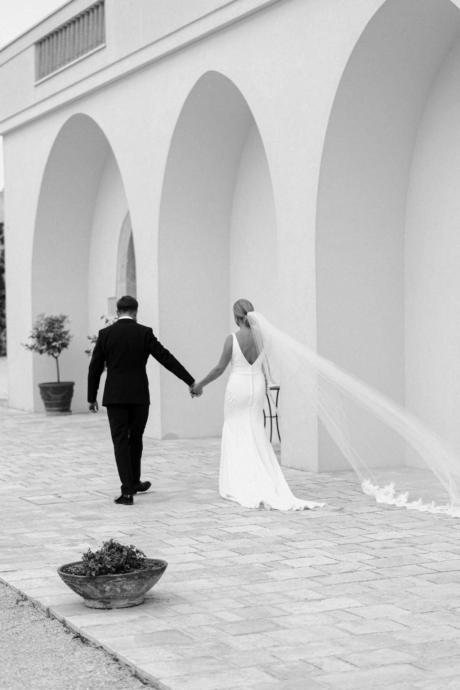 Wedding couple walks through an elegant white archway corridor with Mediterranean architecture.
