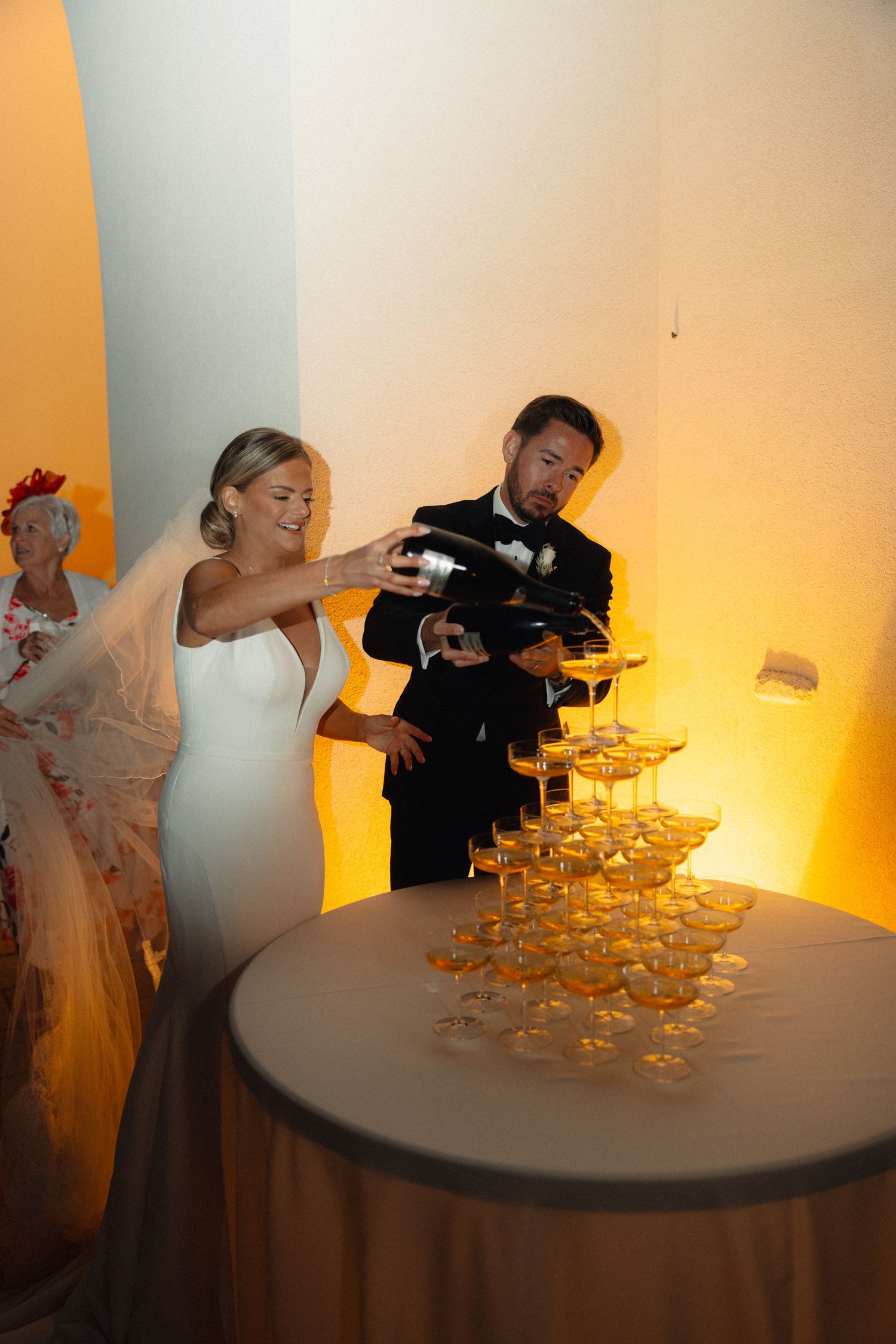 A couple pours champagne into a tower of coupe glasses at an elegant event.