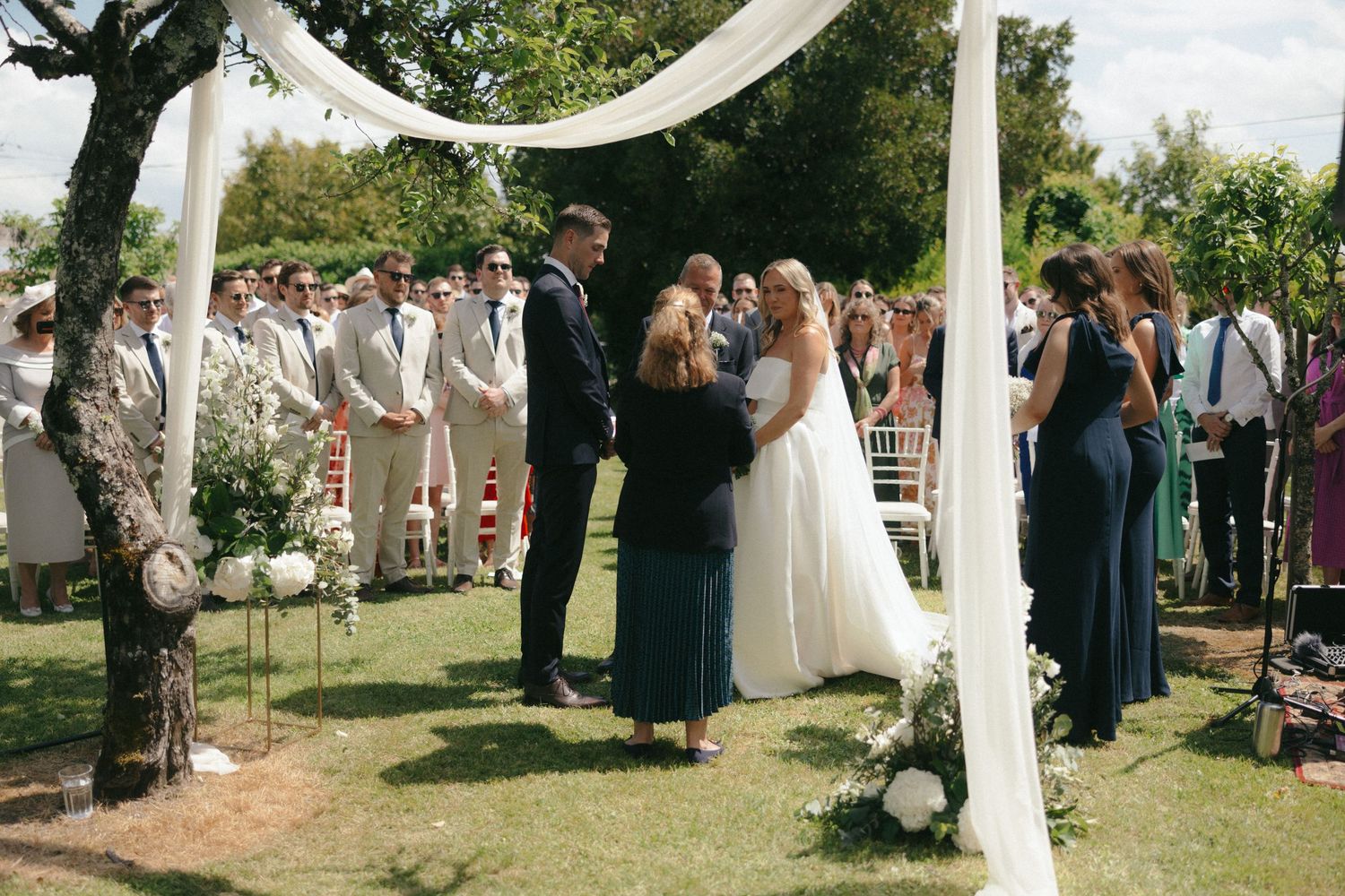 An outdoor wedding ceremony under a white draped arch with guests gathered on a sunny day.