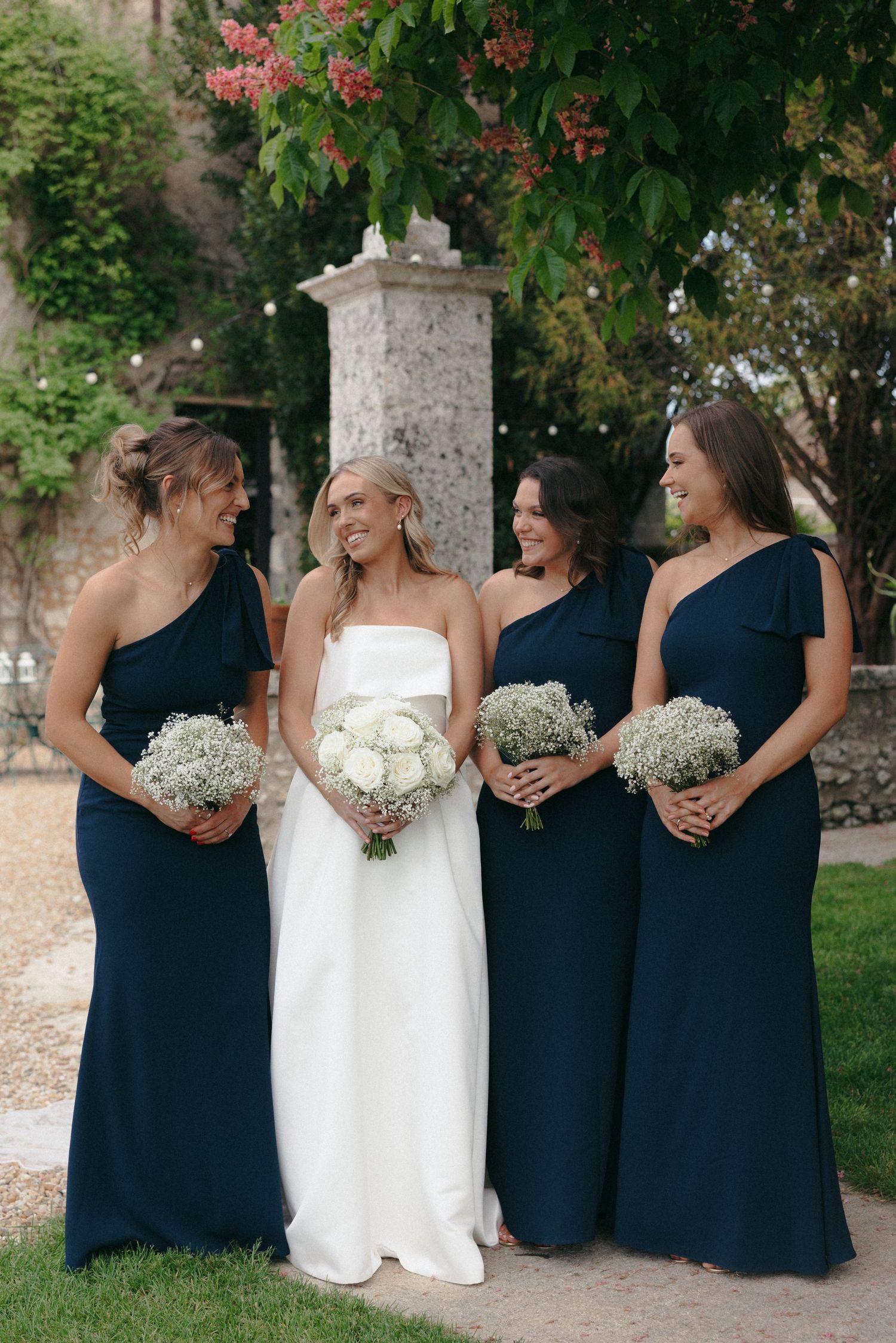 Wedding party poses in navy blue bridesmaid dresses and white bridal gown against outdoor garden setting.