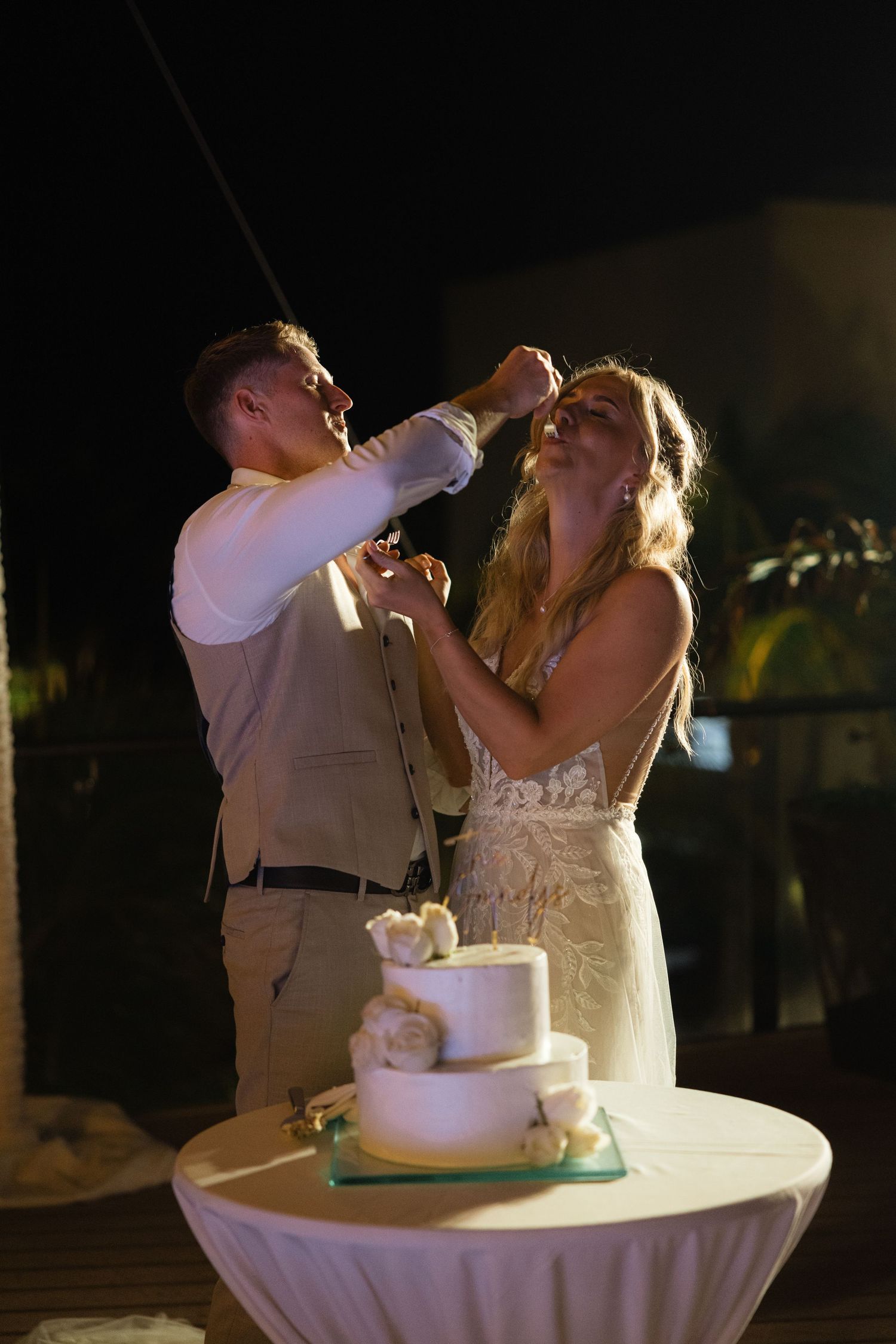 A couple laughs while feeding each other bites of their tiered white wedding cake at their reception.