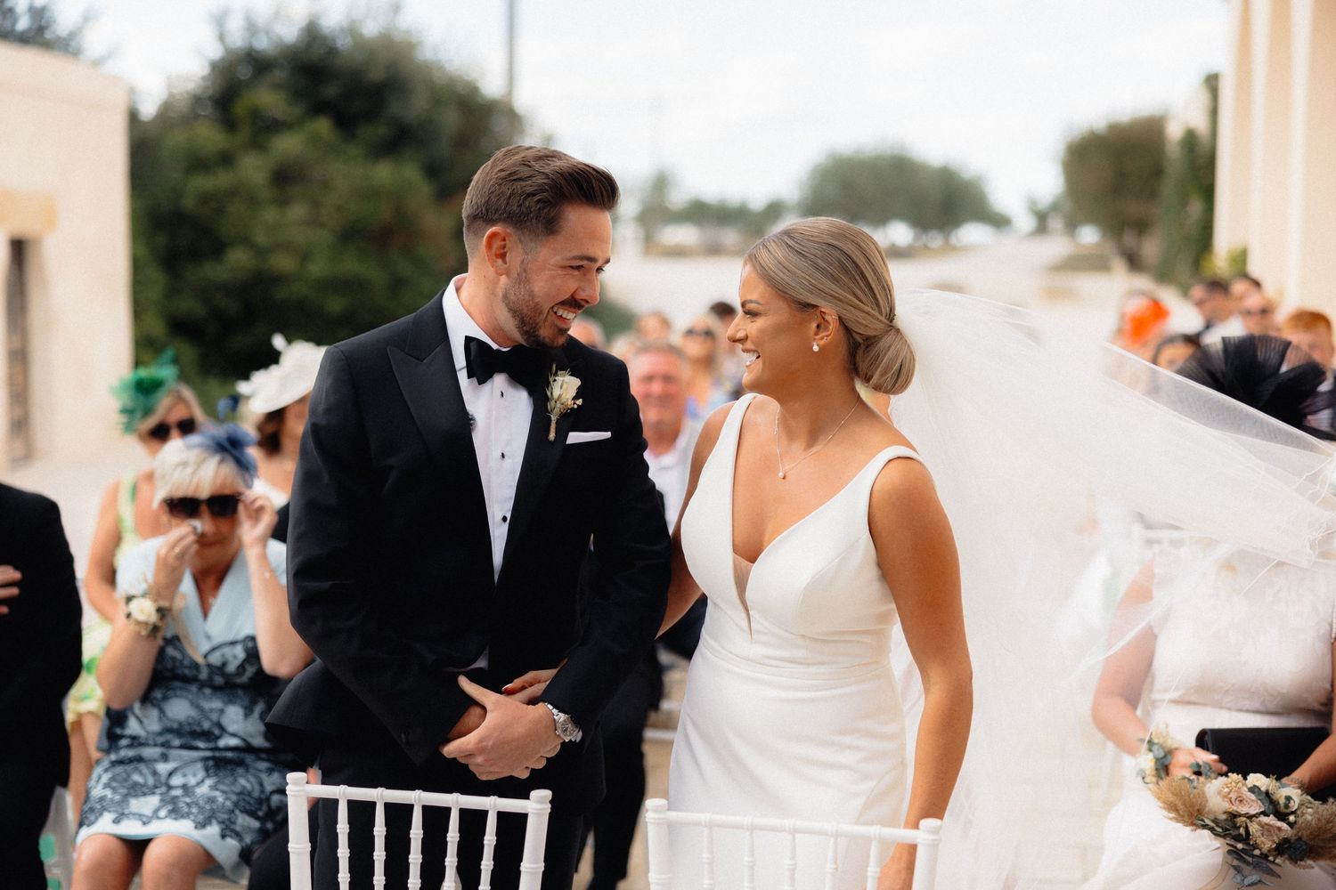 Guests and wedding party members mingle during an outdoor celebration with white chairs.