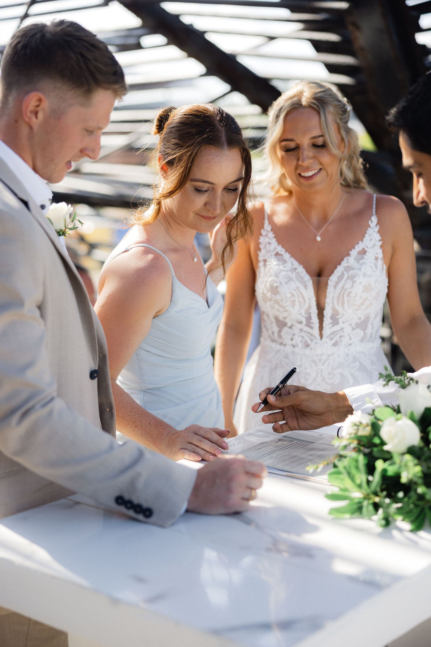 Wedding party signing marriage documents at white table with floral decorations.