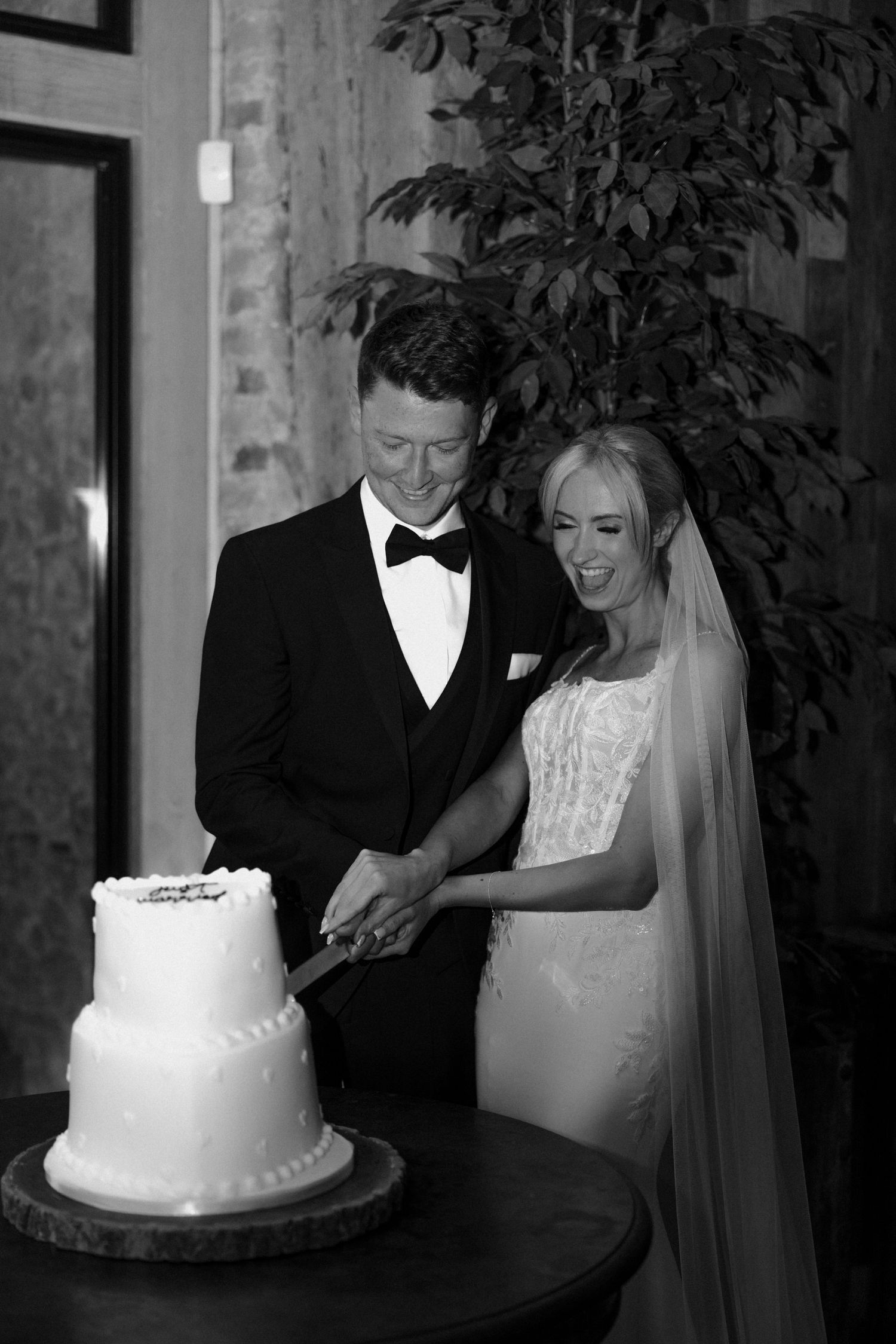 A newlywed couple cuts their elegant two-tier white wedding cake together in black and white photography.
