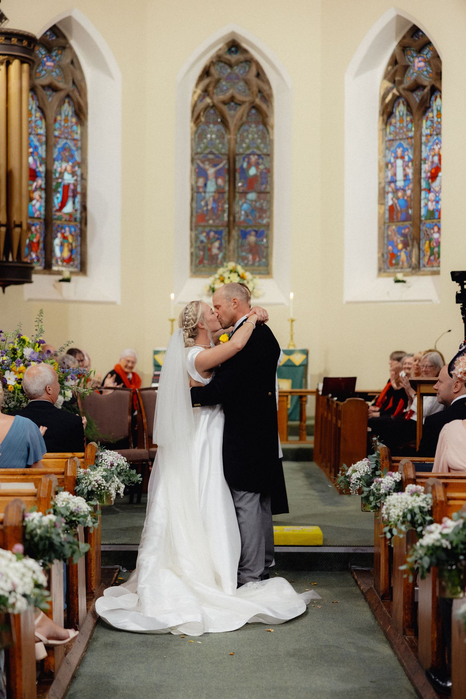 Newlyweds embrace in church aisle decorated with flowers and stained glass windows.