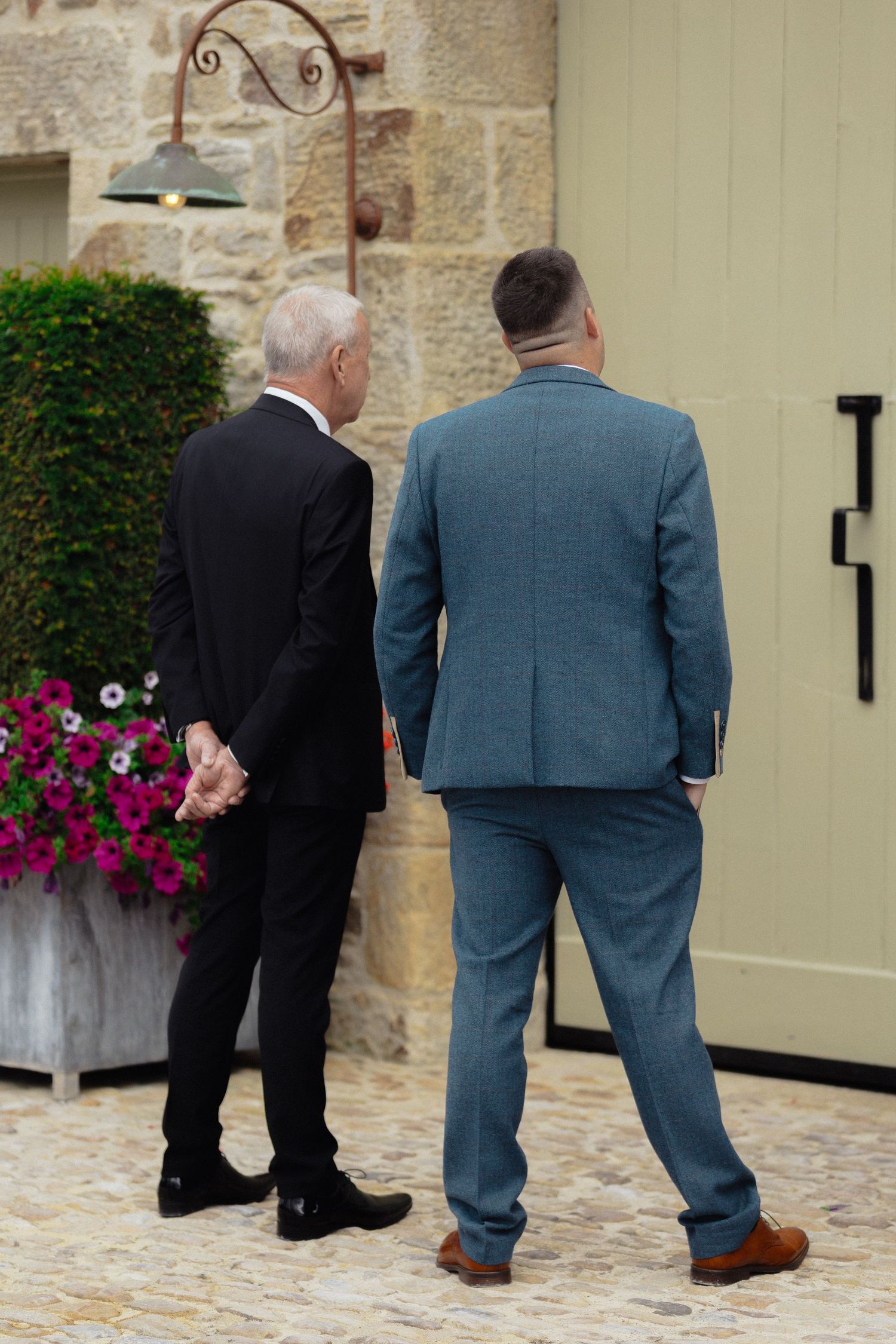 Men in formal suits stand together on a stone patio near colorful flower planters.
