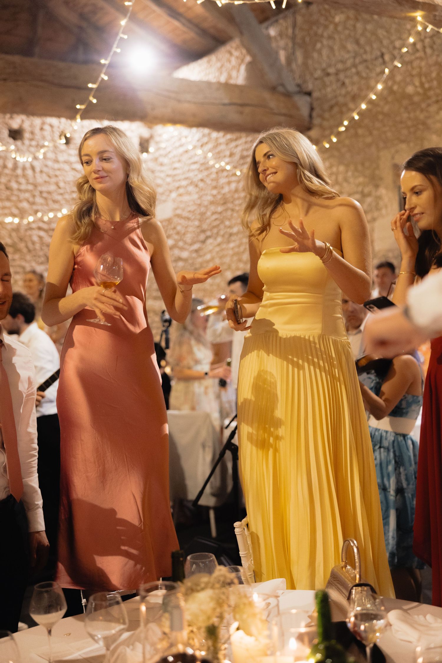 Women in elegant dresses dancing at a wedding reception with string lights and stone walls.