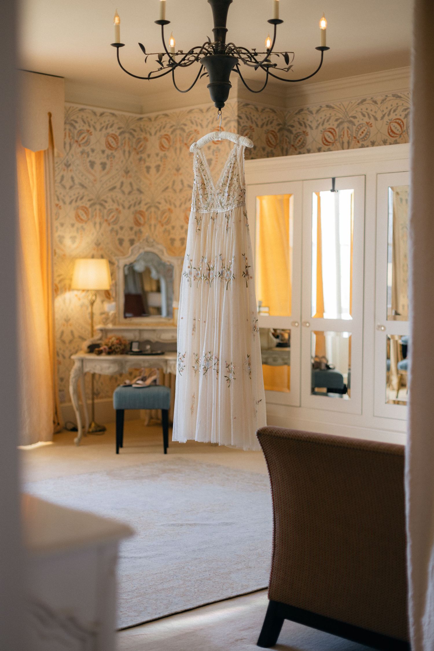 Elegant white lace wedding dress hanging from chandelier in vintage-styled dressing room.