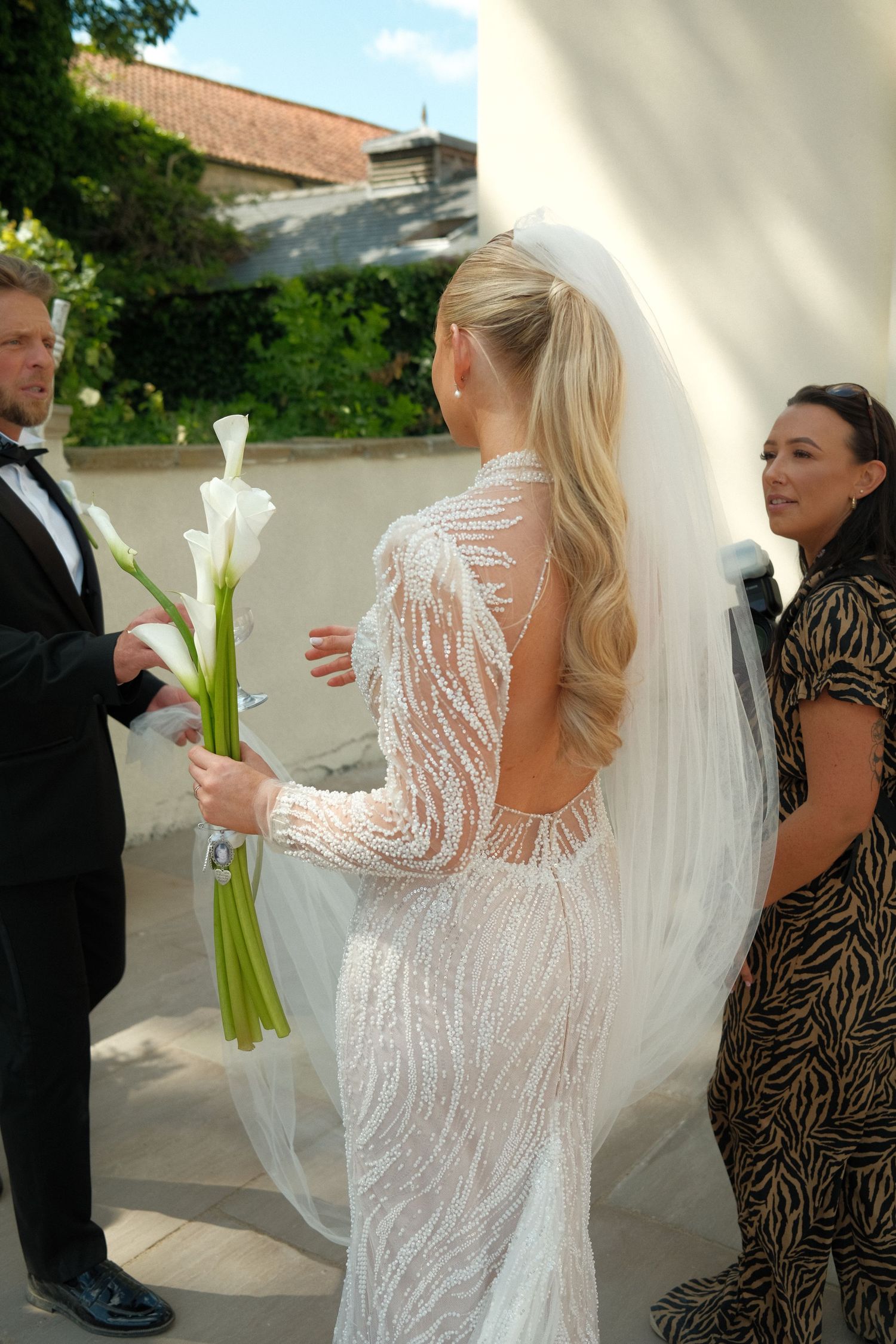 Bride in lace dress holding white calla lilies during outdoor wedding preparations.