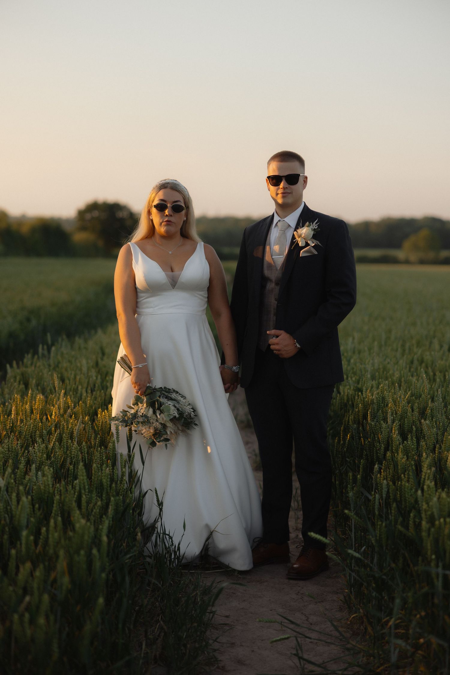 Couple wearing sunglasses walking through a golden wheat field at sunset.
