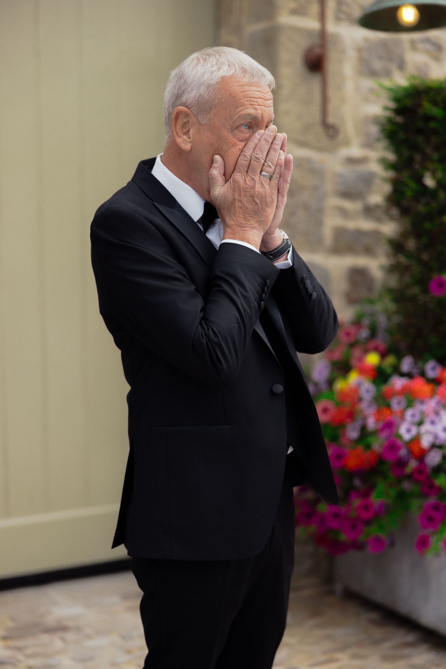 A man in a black tuxedo covers his face with emotion during a wedding ceremony.